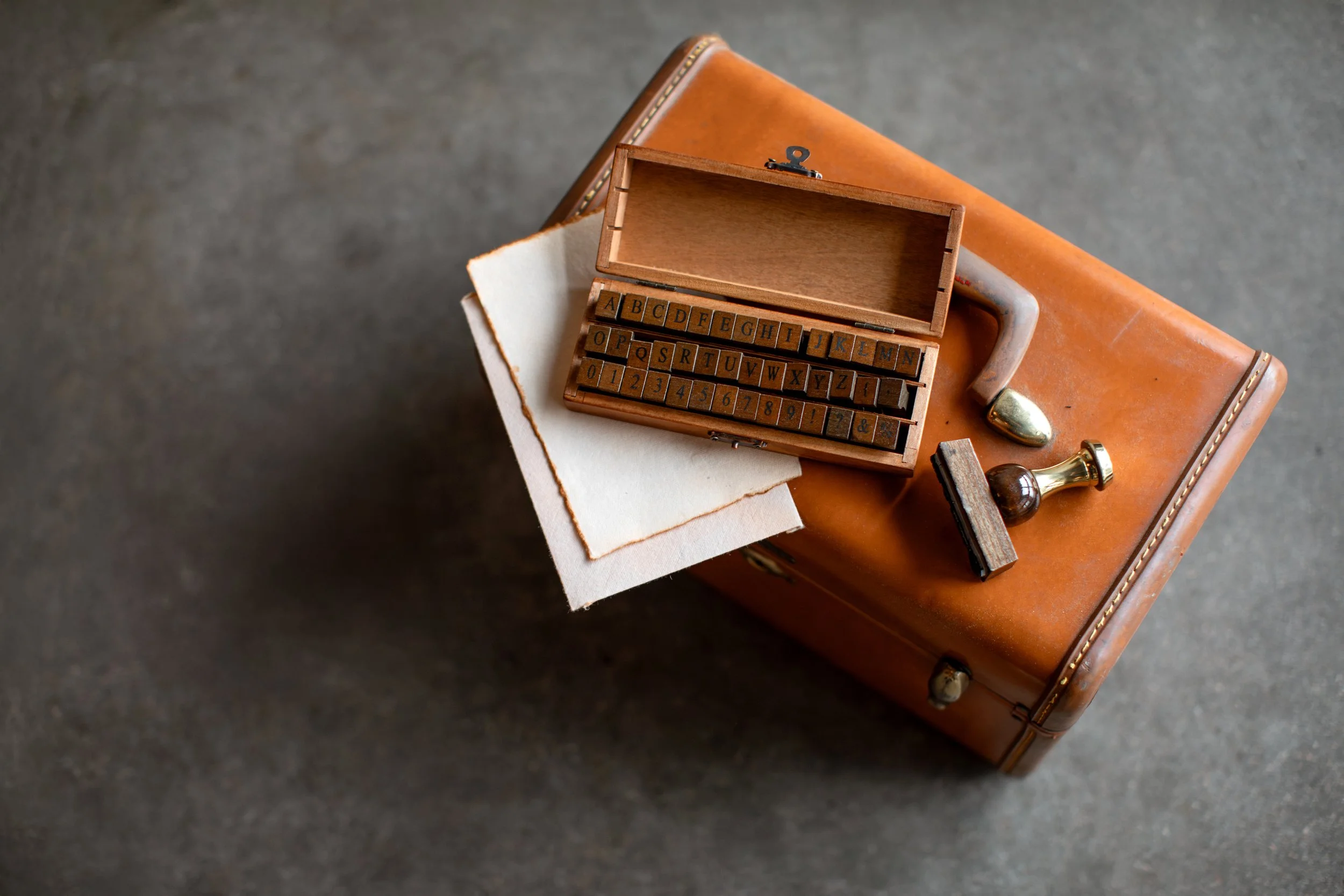 A vintage leather suitcase with a small wooden box containing letterpress type blocks, a piece of paper, and a wax seal stamp on top.
