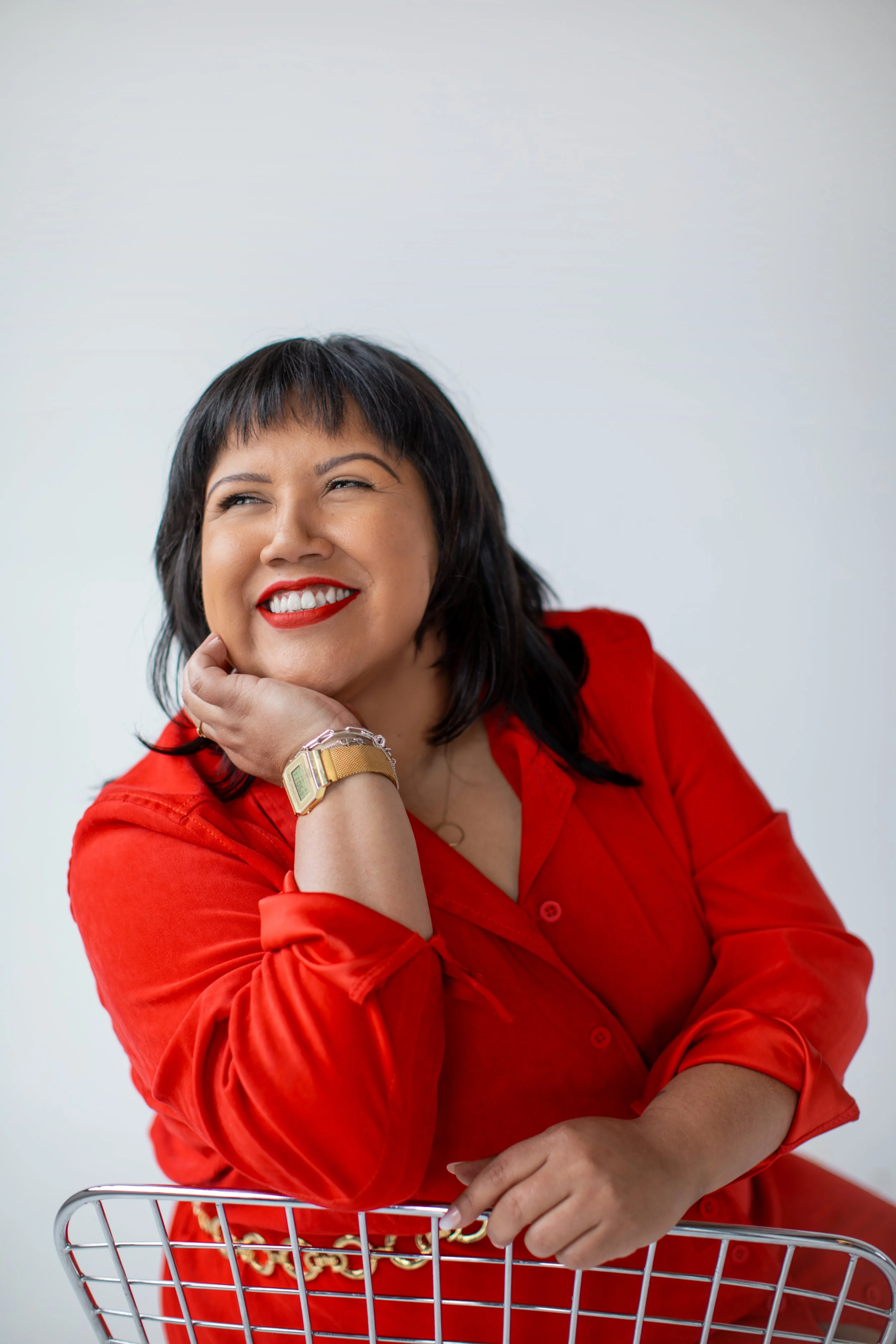 Woman with black hair and red lipstick smiling, wearing a red blouse, sitting behind a shopping cart, with a light gray background.