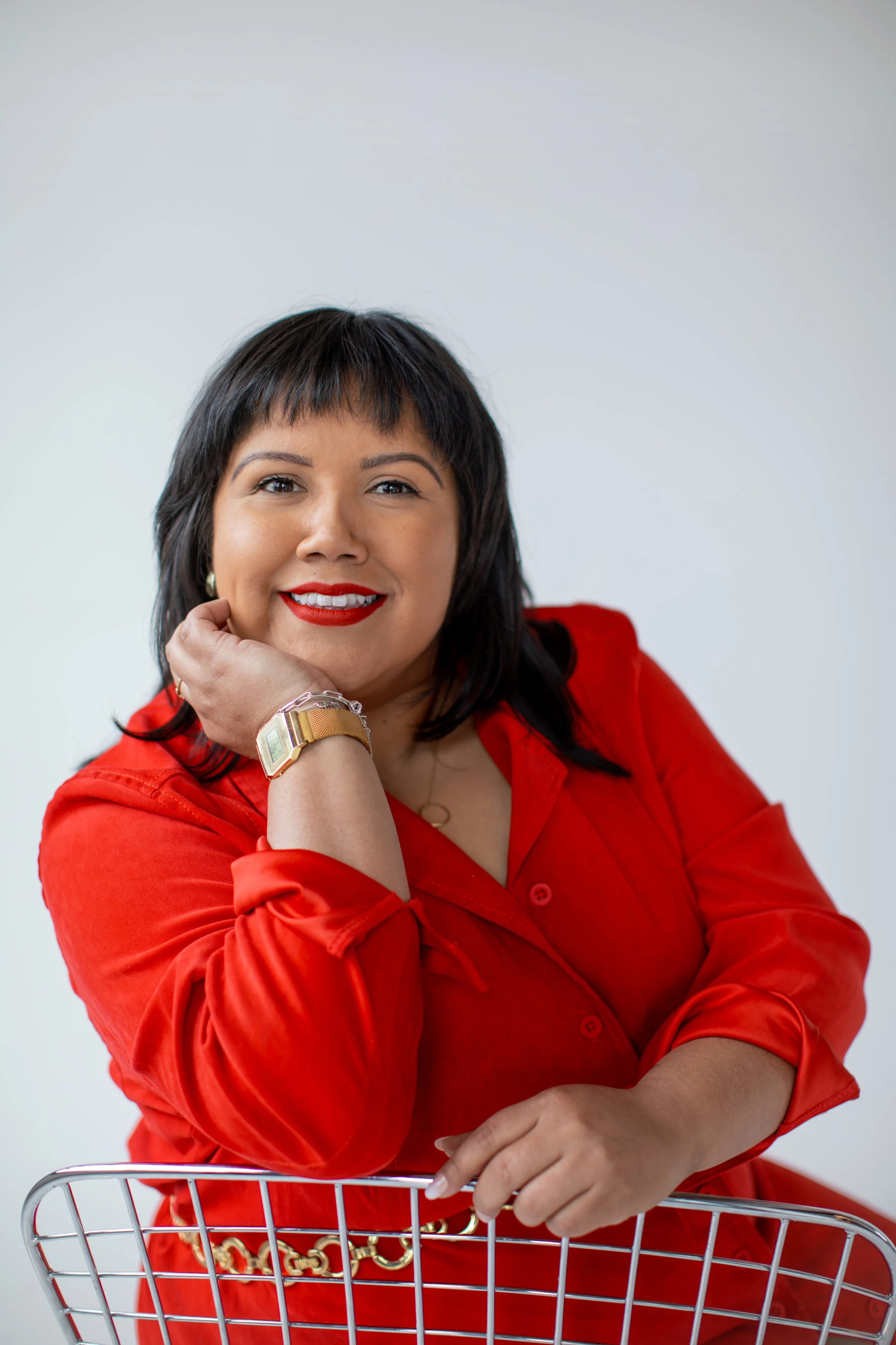 A woman with medium skin tone, dark hair, and red lipstick smiling while resting her chin on her left hand, posing behind a shopping cart, wearing a red blouse and gold accessories, against a plain white background.