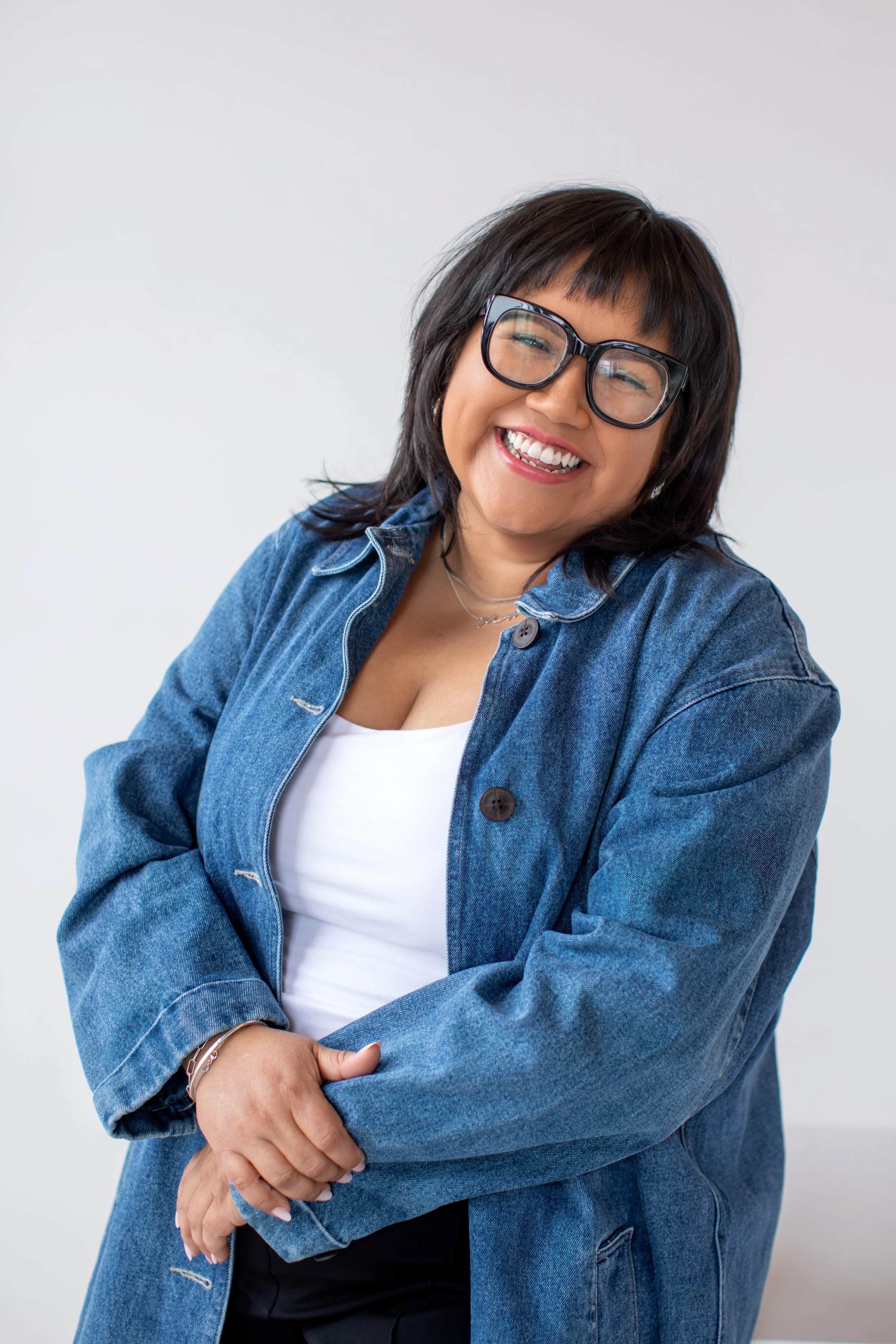 A woman with shoulder-length dark hair, wearing glasses, a white shirt, and a blue denim jacket, smiling and posing with arms crossed against a plain white background.