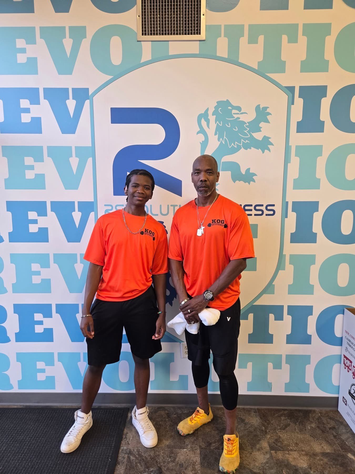 Two men standing in front of a wall with a large logo of a lion and the words 'REVOLUTION' and 'BUSINESS'. Both are wearing orange shirts with 'KOG' printed on them, and are dressed in athletic clothing.