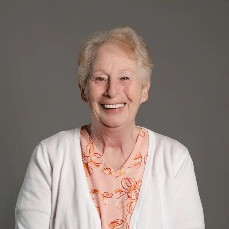 A smiling elderly woman with short, light-colored hair wearing a white cardigan over a peach blouse with floral patterns, against a plain gray background.