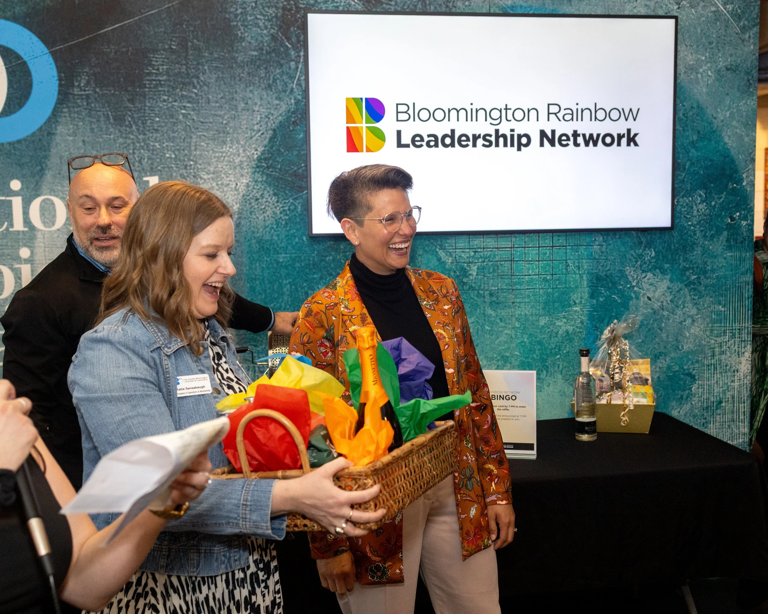 Group of smiling women and a man at a Blooming Rainbow Leadership Network event, with a display screen in the background showing the organization’s logo.