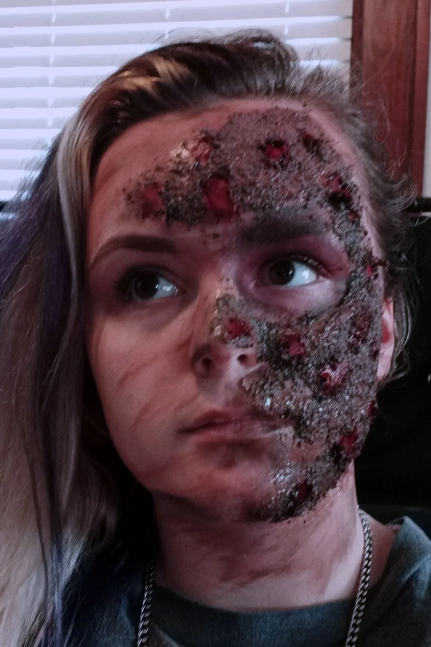 Person with long brown hair lying down with face covered in a dark, gritty substance with redness.