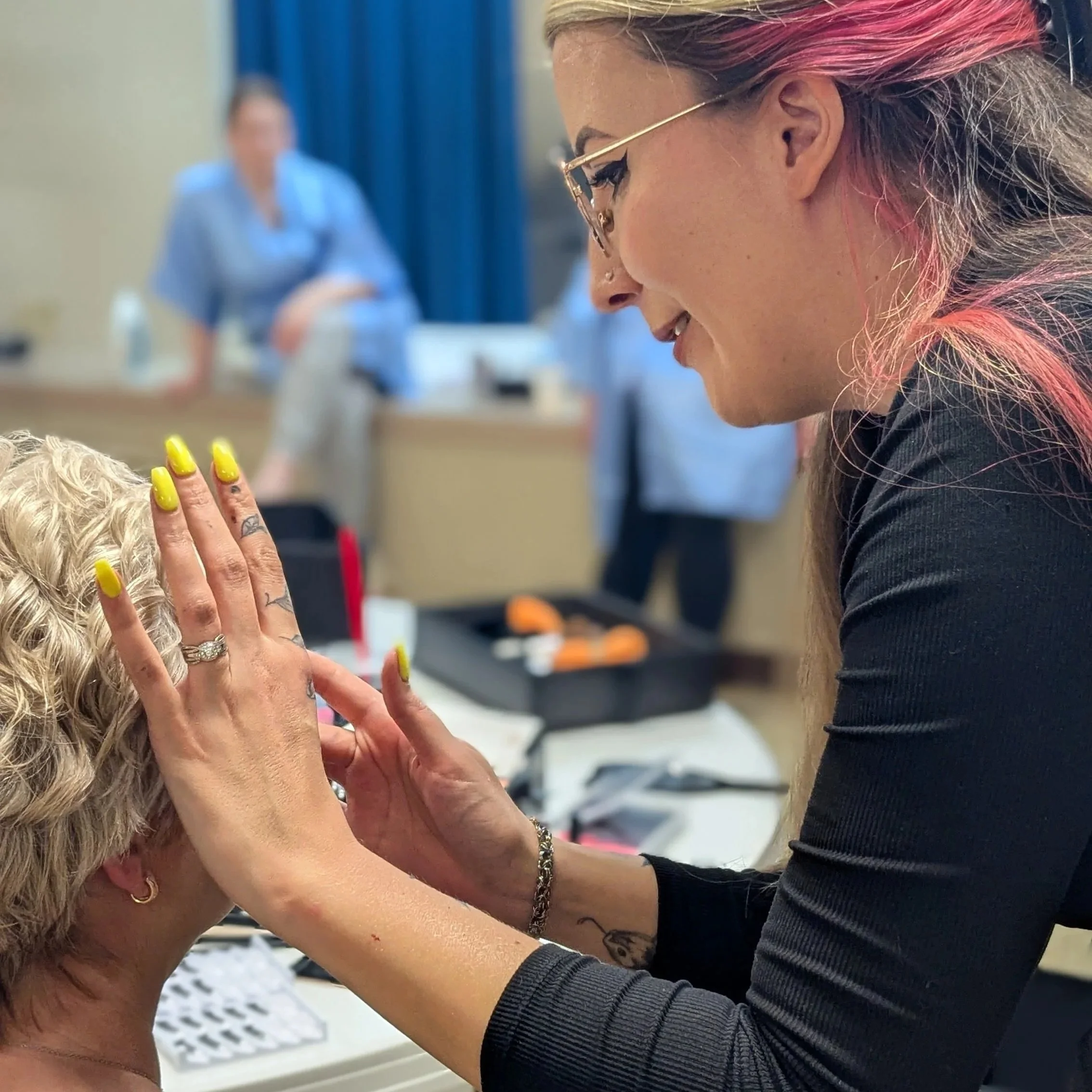 A tattooed woman wearing glasses with pink highlights on her hair, smiling and touching someone's head with long blonde hair and vibrant yellow nails, in an indoor setting with a blurred person in a blue shirt in the background.