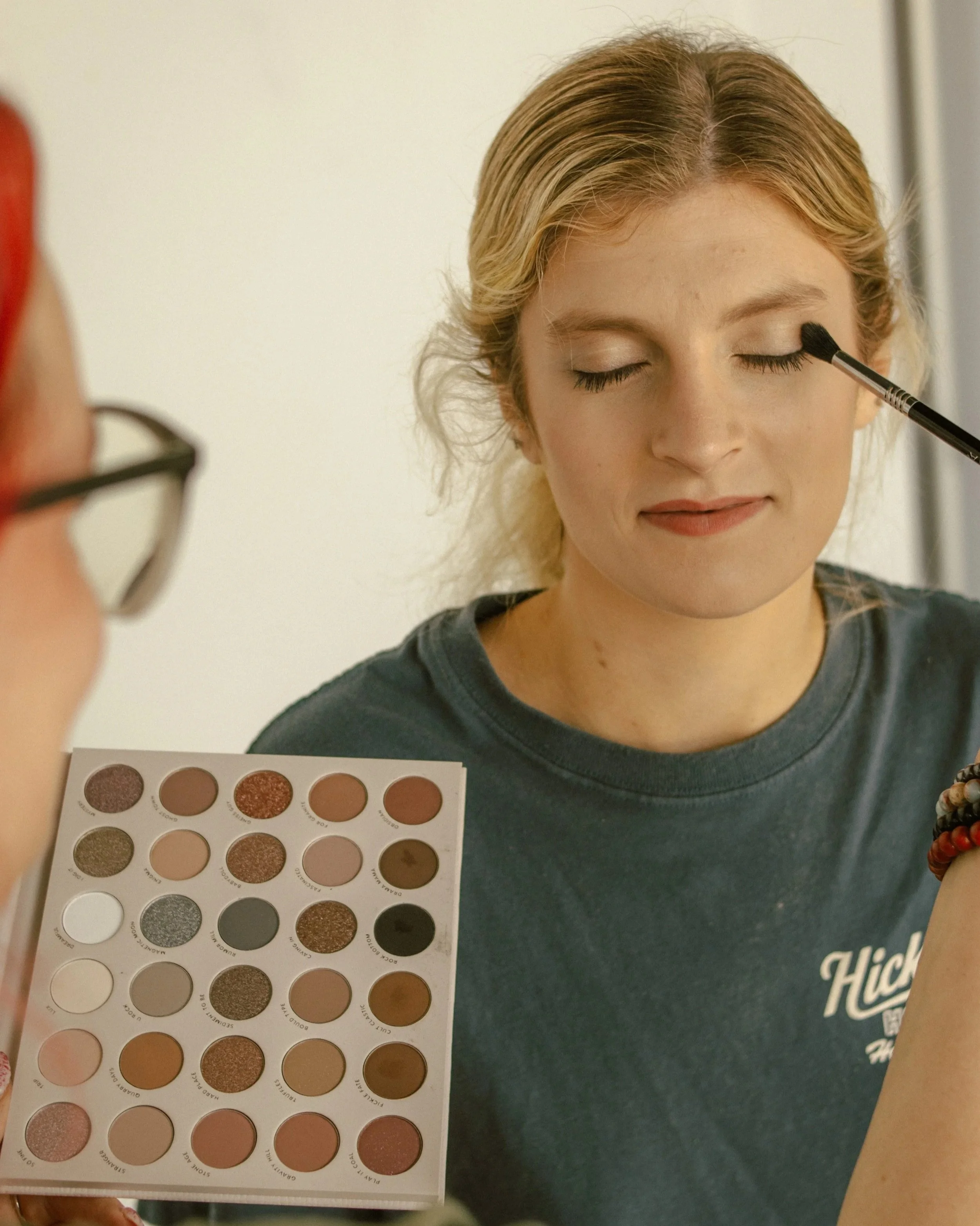 A woman with blonde hair applies eye makeup using a black brush, with a makeup palette of eye shadows in various neutral and shimmery shades on a white rectangular palette in front of her.