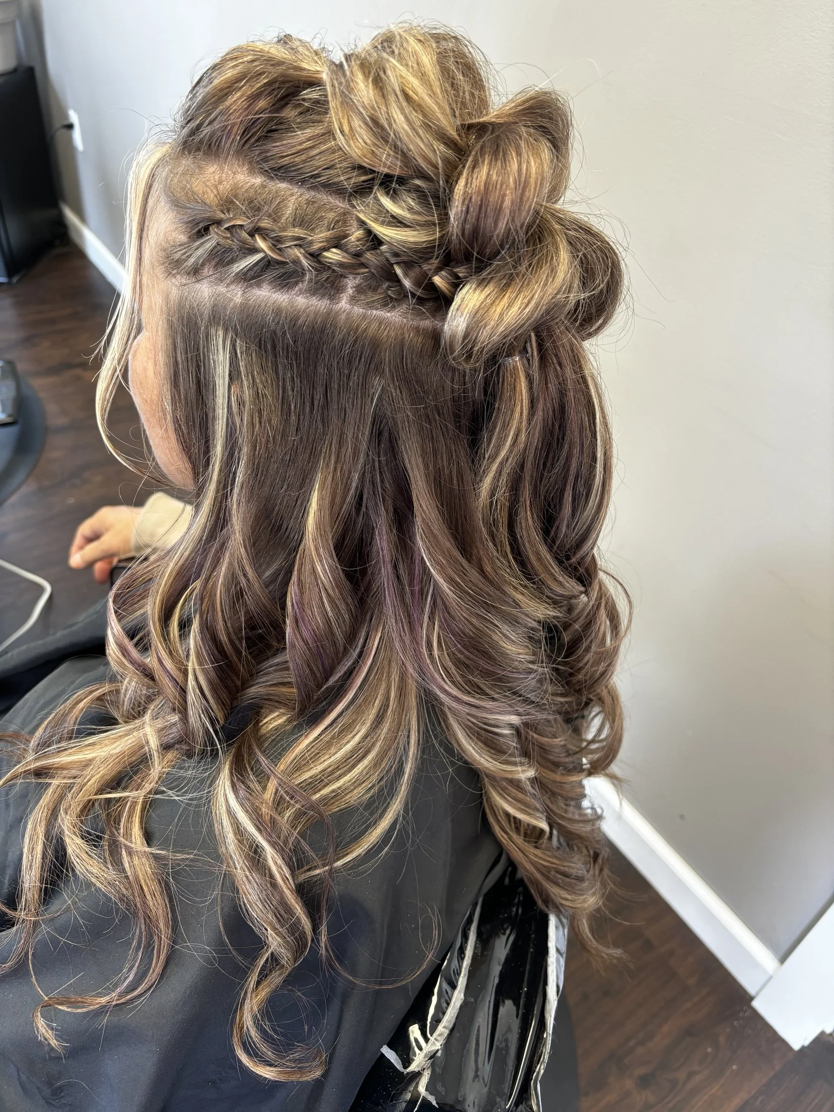 A woman with multi-colored hair styled with loose curls and a small braid on the side, sitting in a salon chair with a cape around her shoulders.