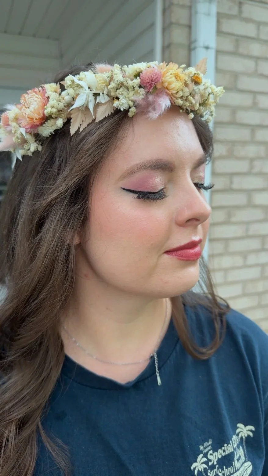 A young woman with long brown hair wearing a floral crown and makeup with winged eyeliner, pink eyeshadow, and pink lipstick, standing outdoors in front of a brick wall.