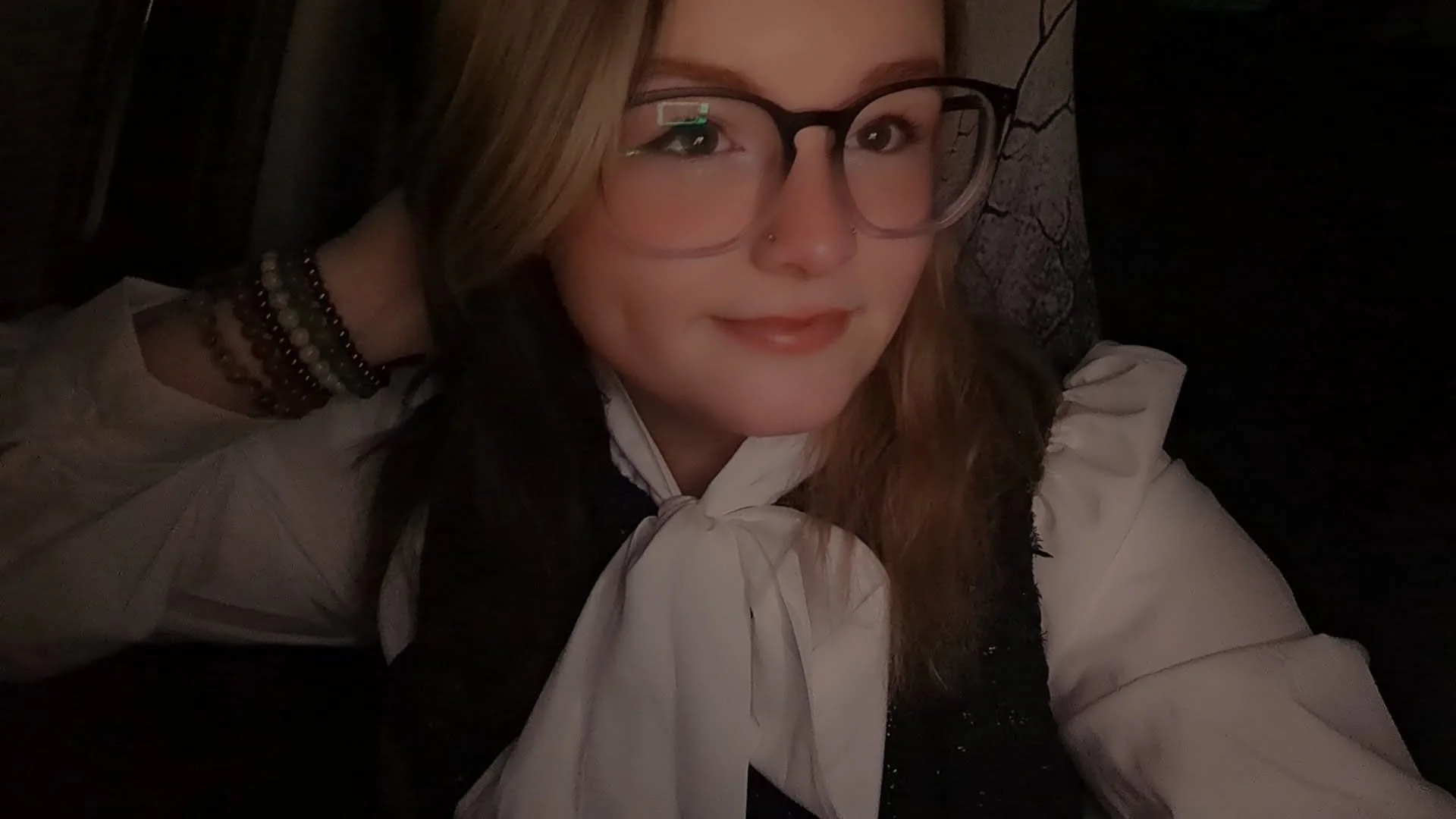 A woman with long reddish hair, wearing glasses, a white blouse with a bow, and jewelry, smiling softly in a dimly lit setting.