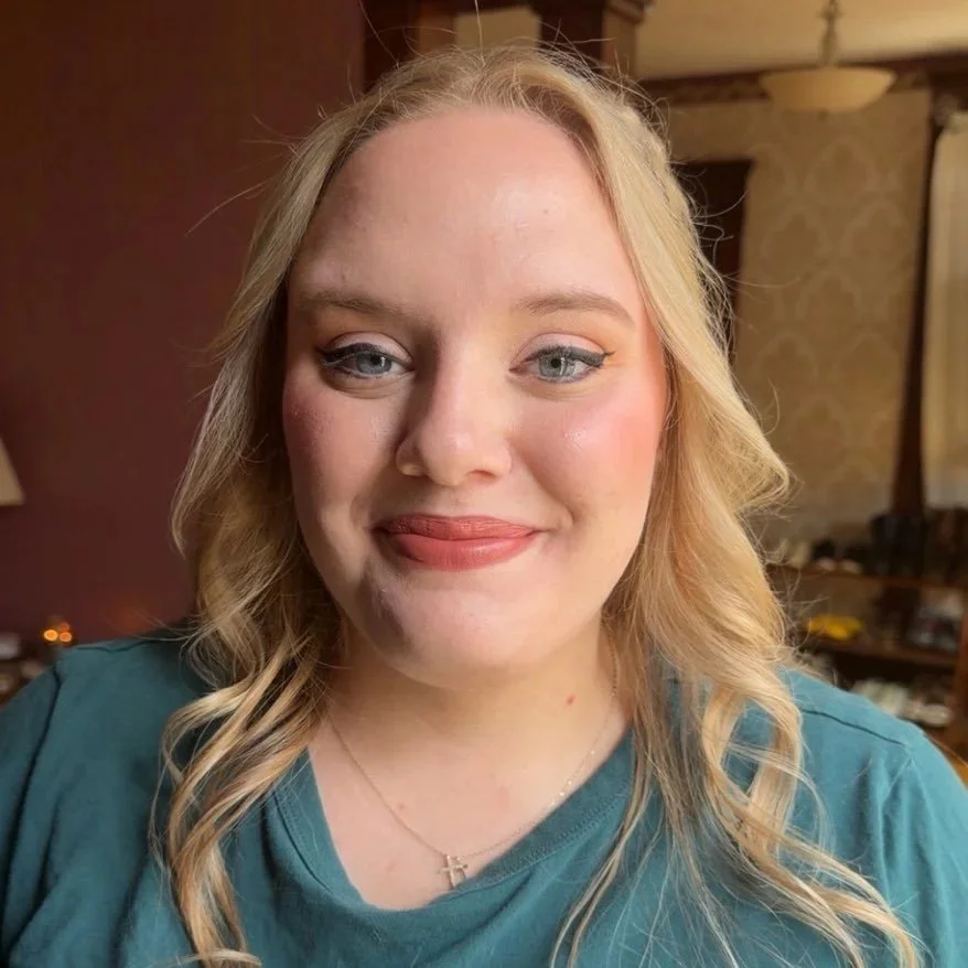 A woman with blonde hair, blue eyes, and light makeup smiling in a room with warm lighting and patterned wallpaper.