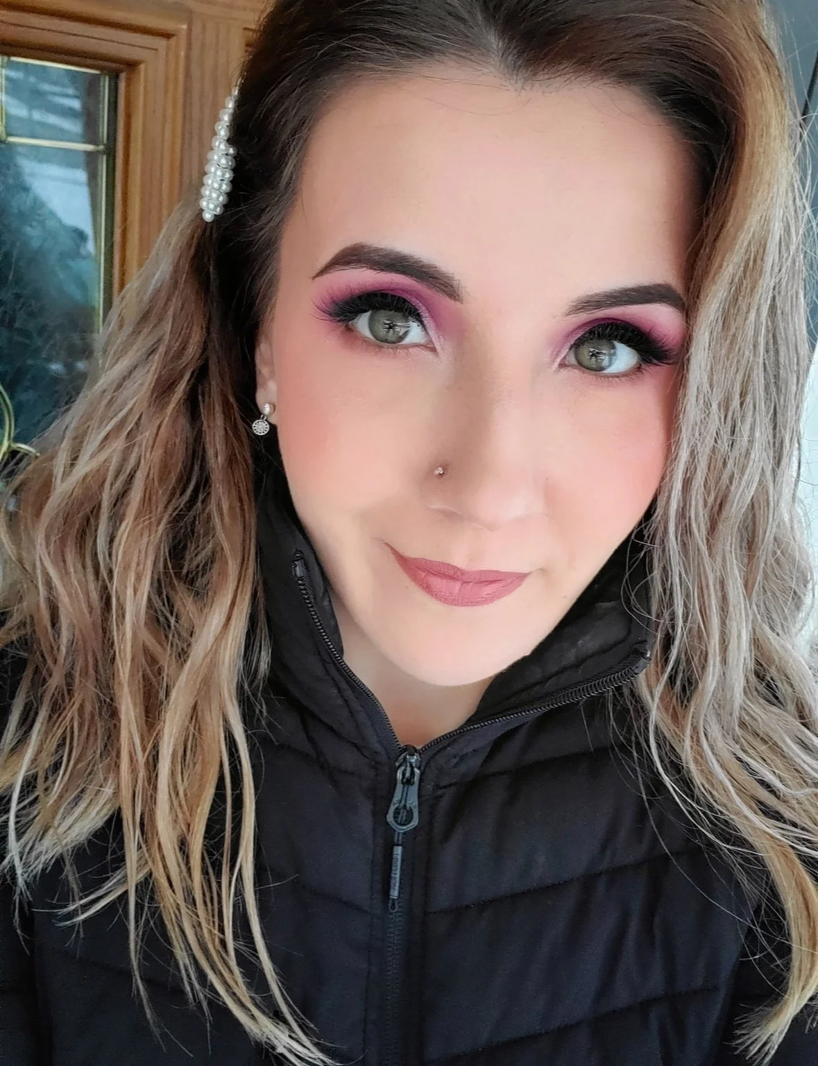 Close-up of a woman with long wavy blonde hair, wearing makeup with pink eyeshadow, light lipstick, a nose piercing, pearl earrings, and a black zip-up jacket, standing in front of a wooden door.