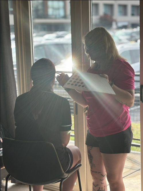 A makeup artist applying makeup to a woman sitting on a chair near a glass door during daytime.