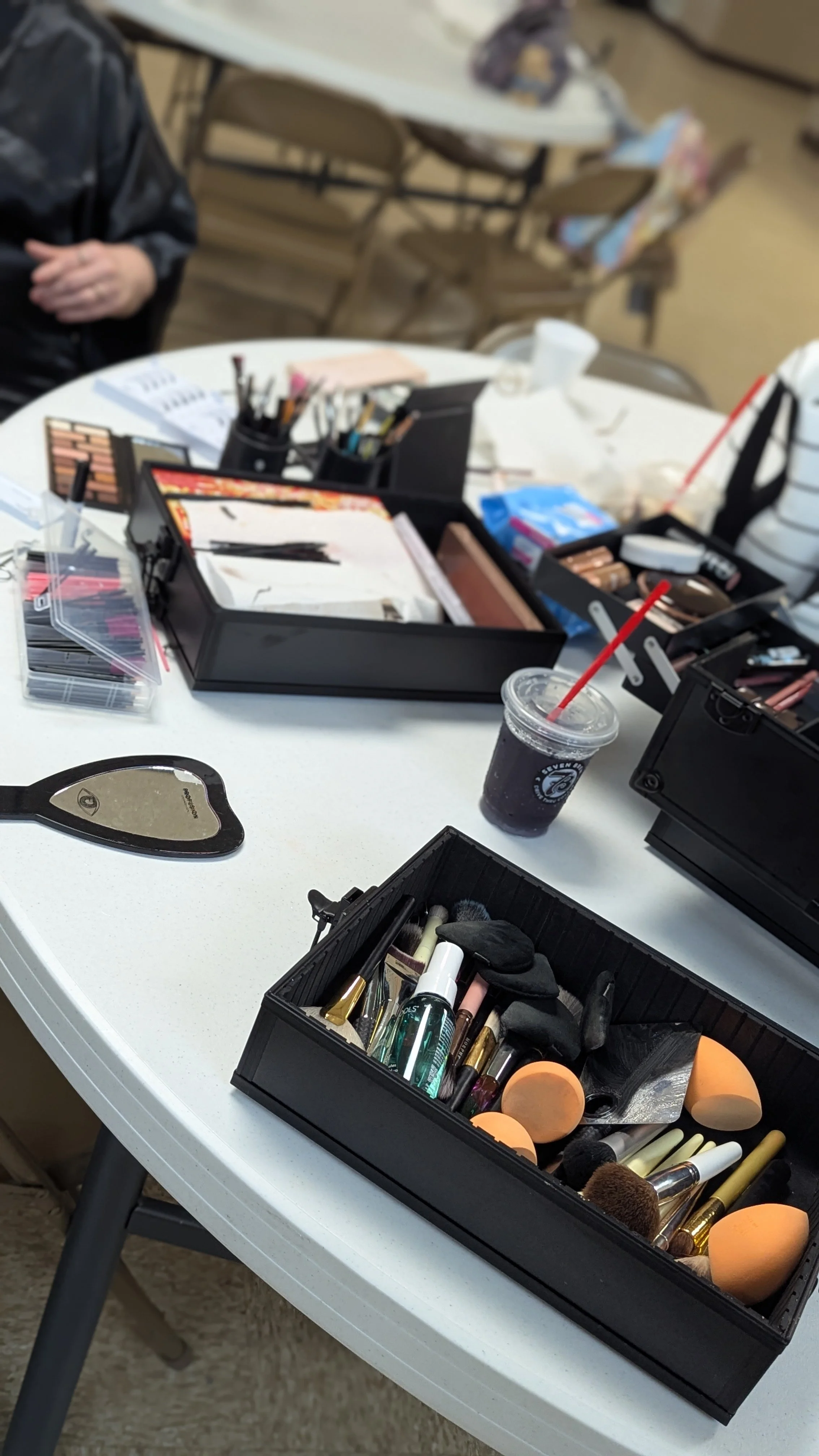 Makeup tools and products on a white round table, including brushes, sponges, lip gloss, eyeshadow palette, and a cold beverage with a straw.