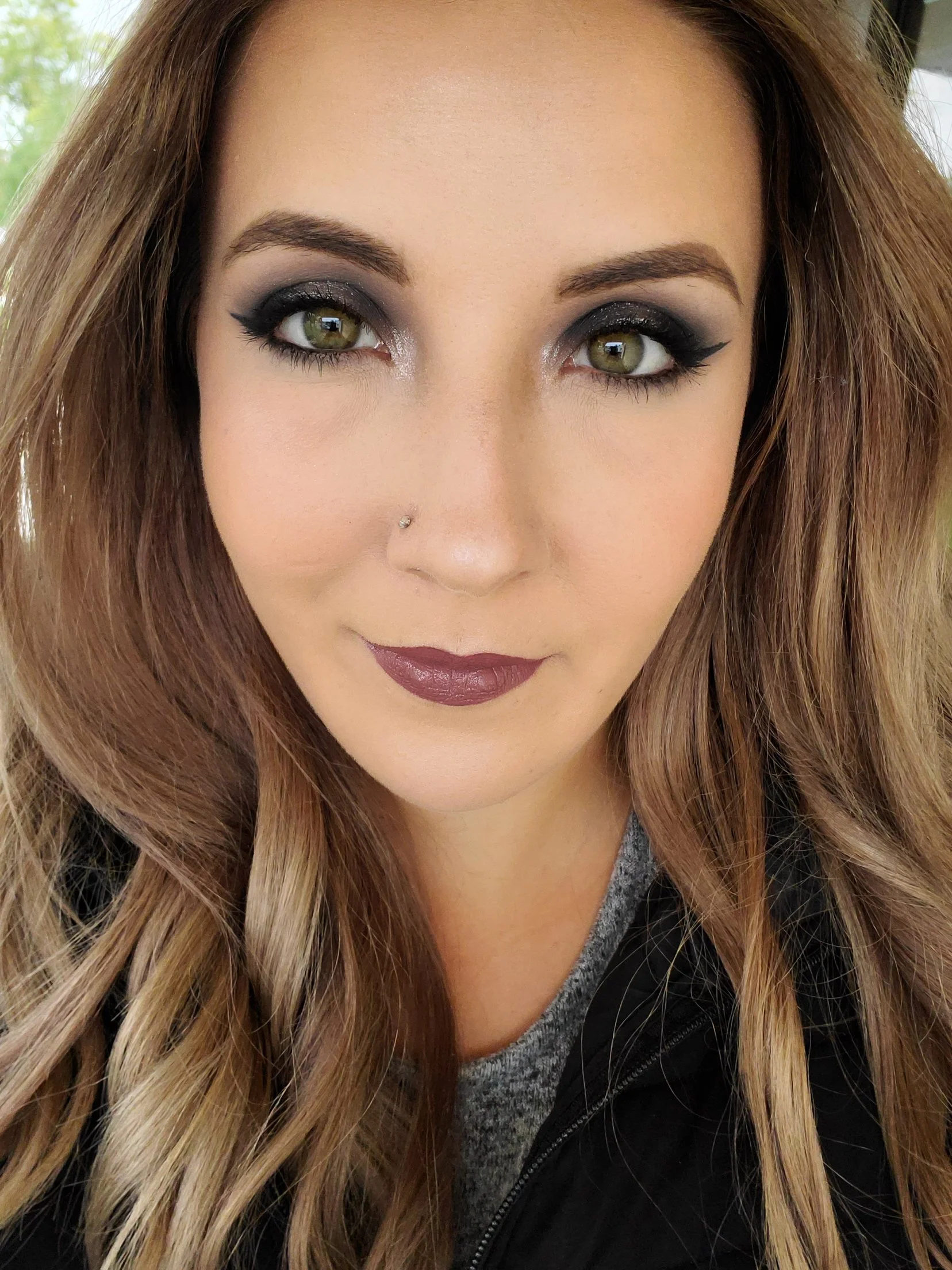 Close-up of a woman with long, wavy hair, green eyes, dark eye makeup, and mauve lipstick, wearing a gray shirt and black jacket.