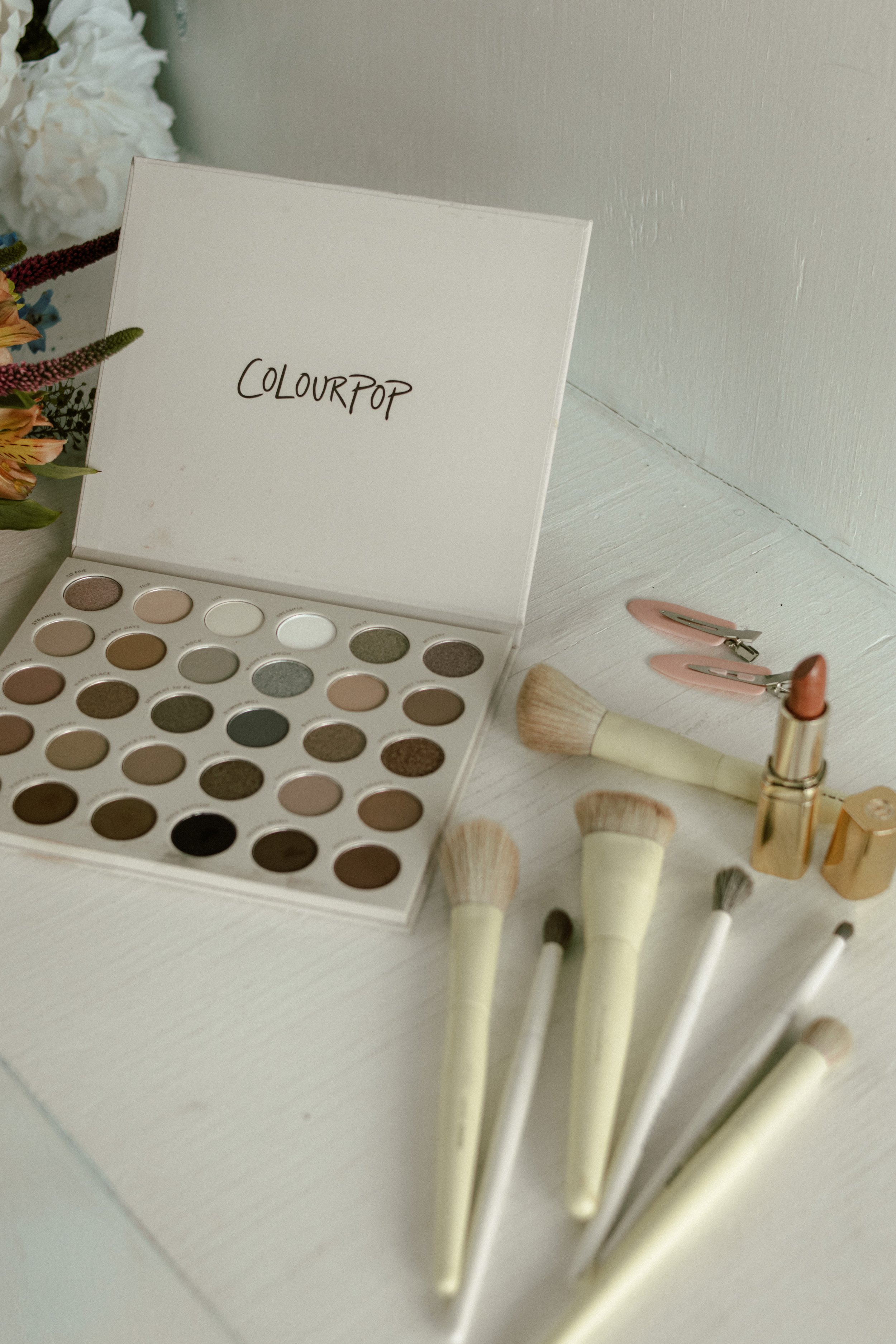 A white eyeshadow palette with various shades, some matte and some shimmer, next to makeup brushes and pink hair clips on a white surface.