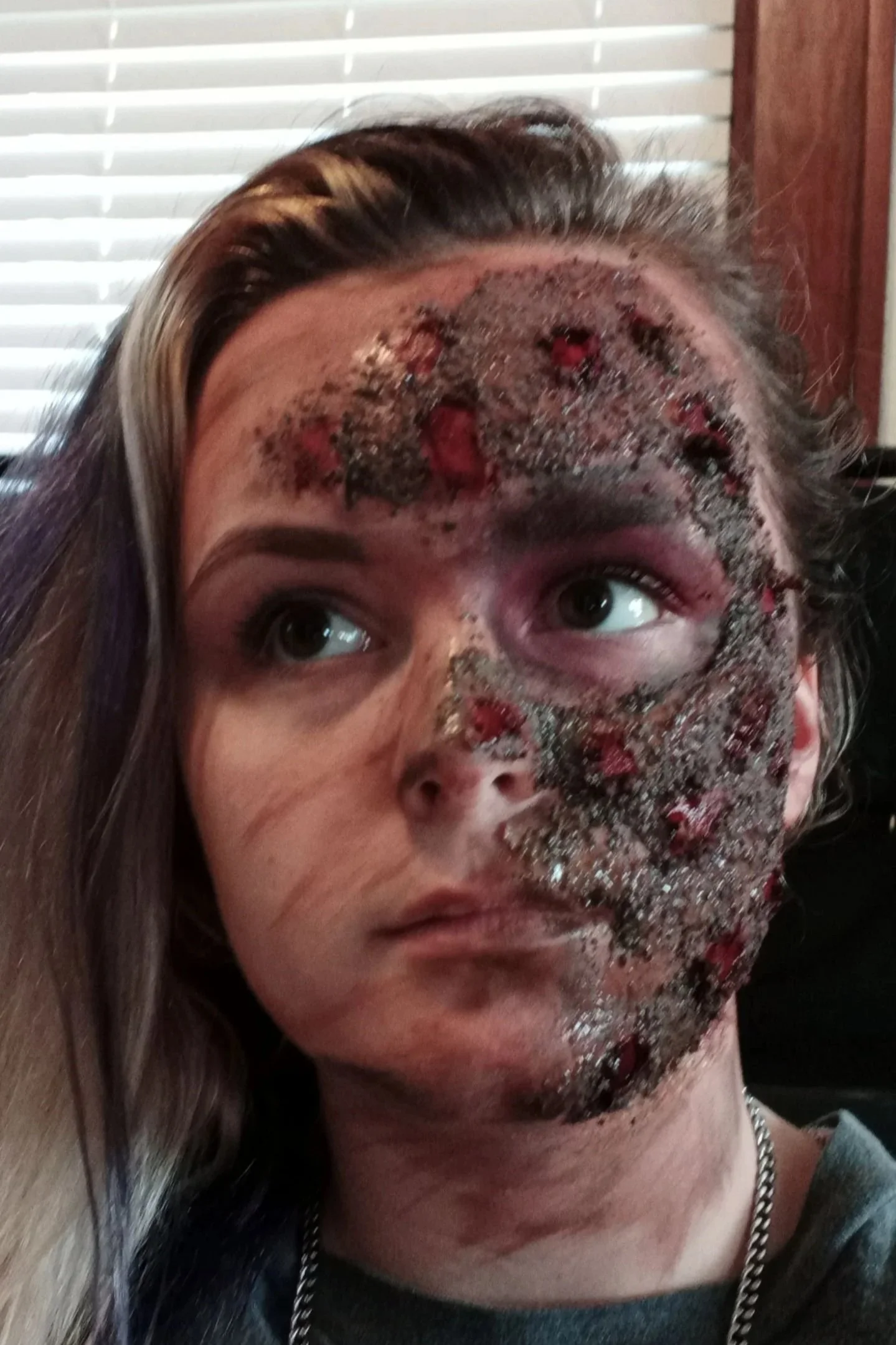 A woman lying on her side with her head resting on a cushion, her face covered with a facial mask with speckles and red spots, and her hair tucked behind her ear.
