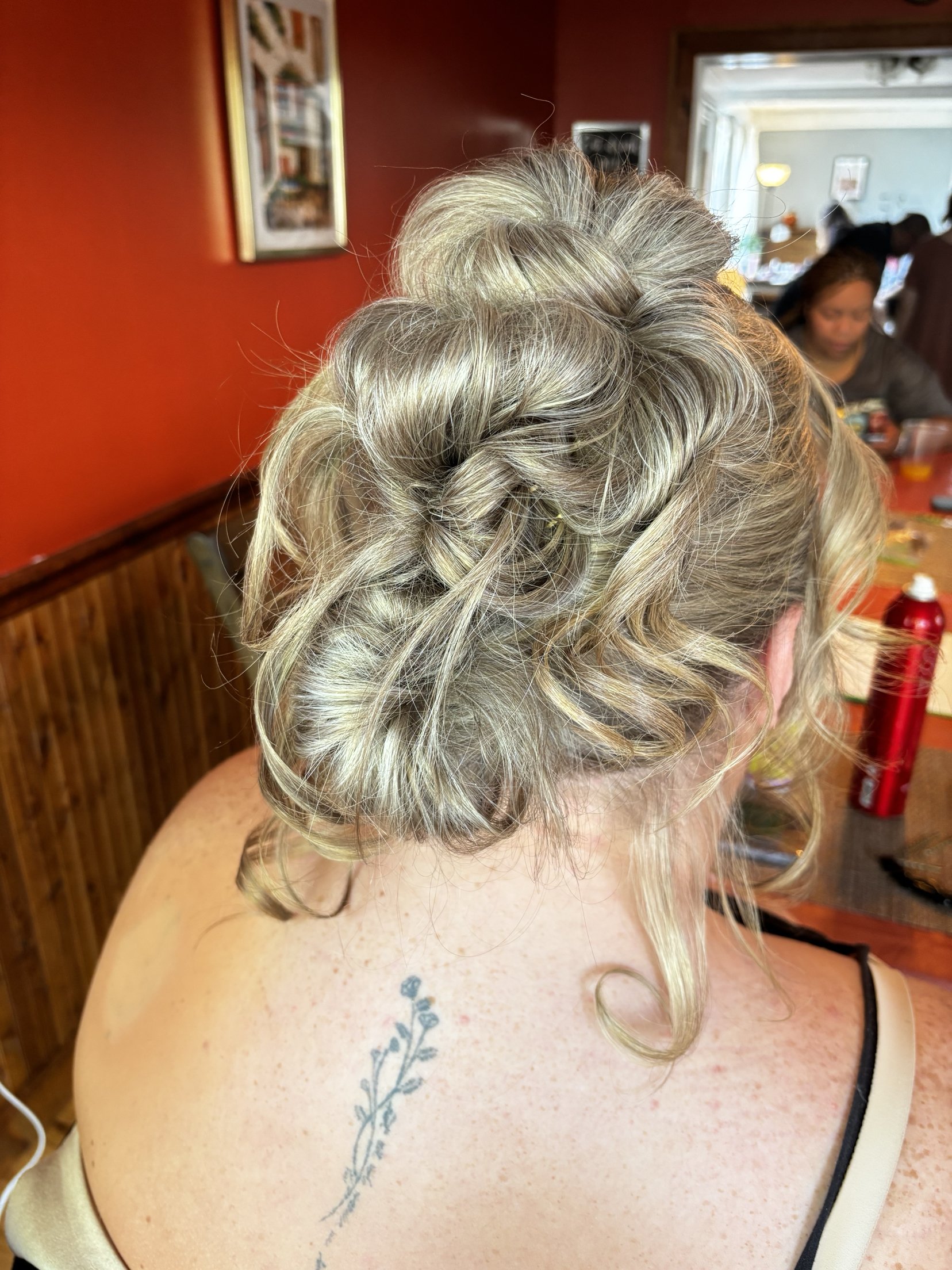 A woman with curly blonde hair styled in an updo, seated at a table with a floral tattoo on her shoulder and back, in a restaurant setting.