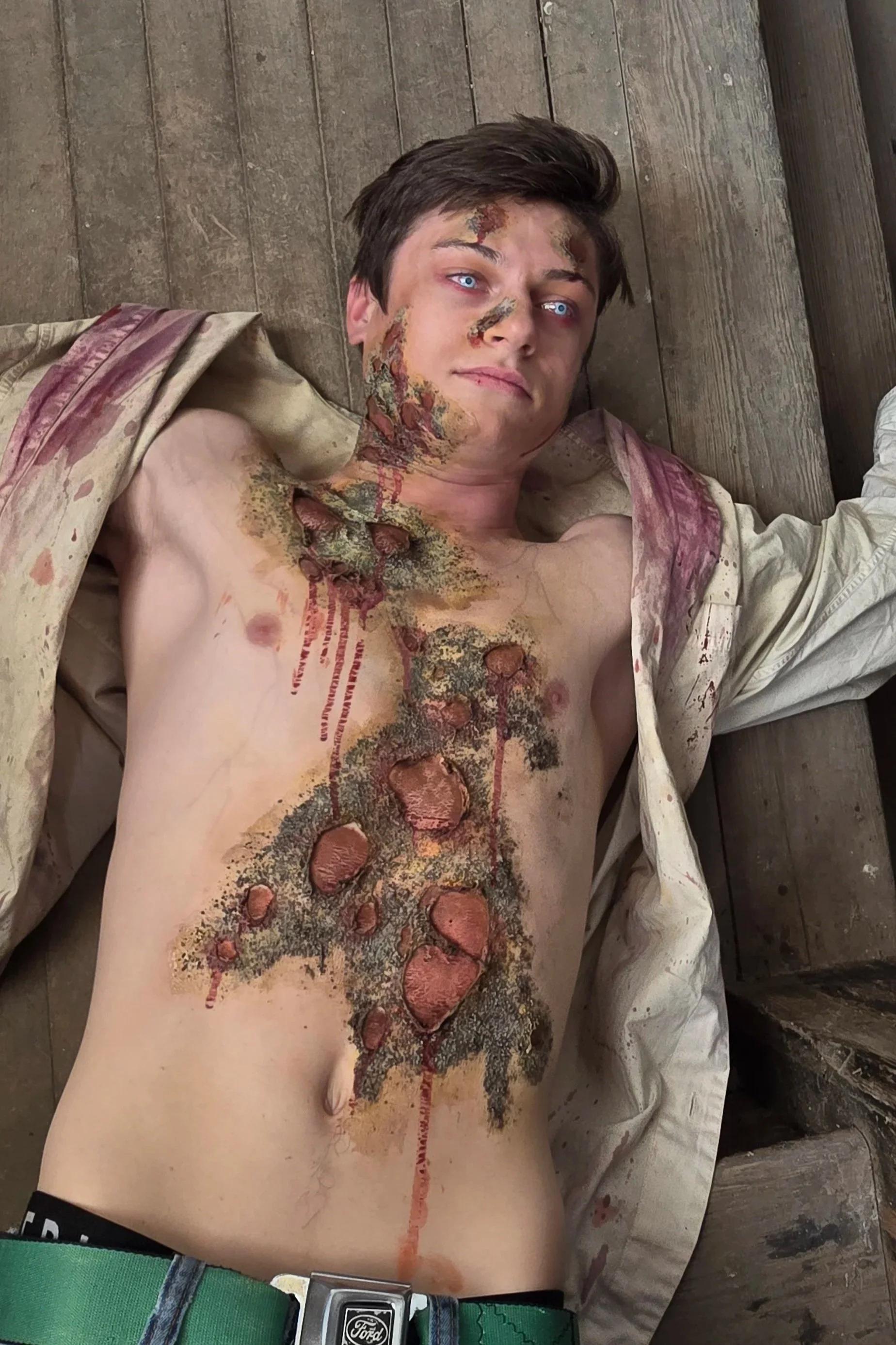 A young man with blue eyes lying on a wooden floor, covered in simulated injuries and blood, wearing a beige shirt, green belt, and gray pants.