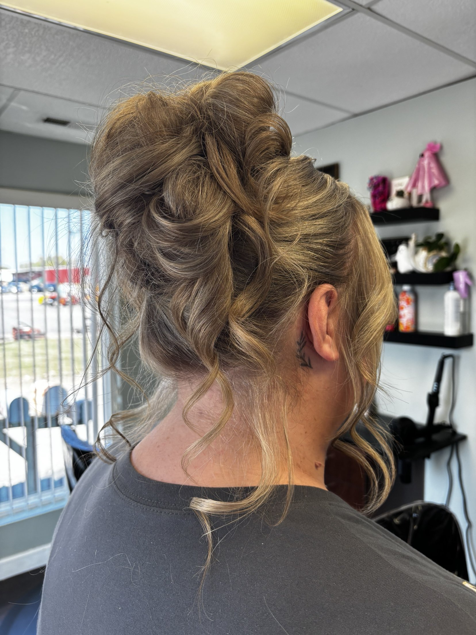Close-up of a woman with styled blonde hair, showing intricate curls and an updo, inside a salon with a large window in the background.