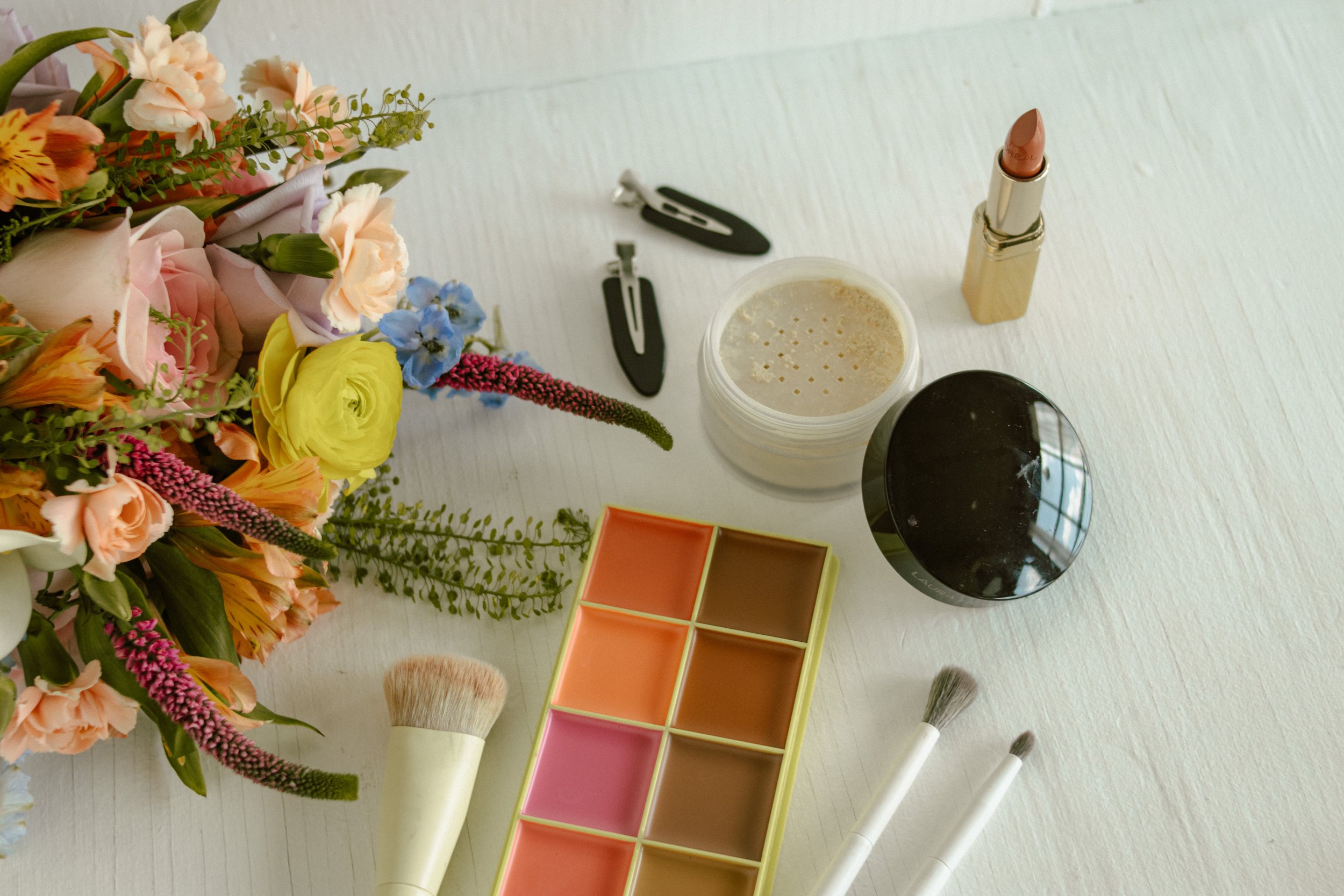 A flat lay of makeup items including lipstick, makeup brushes, an eyeshadow palette, a container of loose powder, a mirror, and a flower bouquet on a white surface.