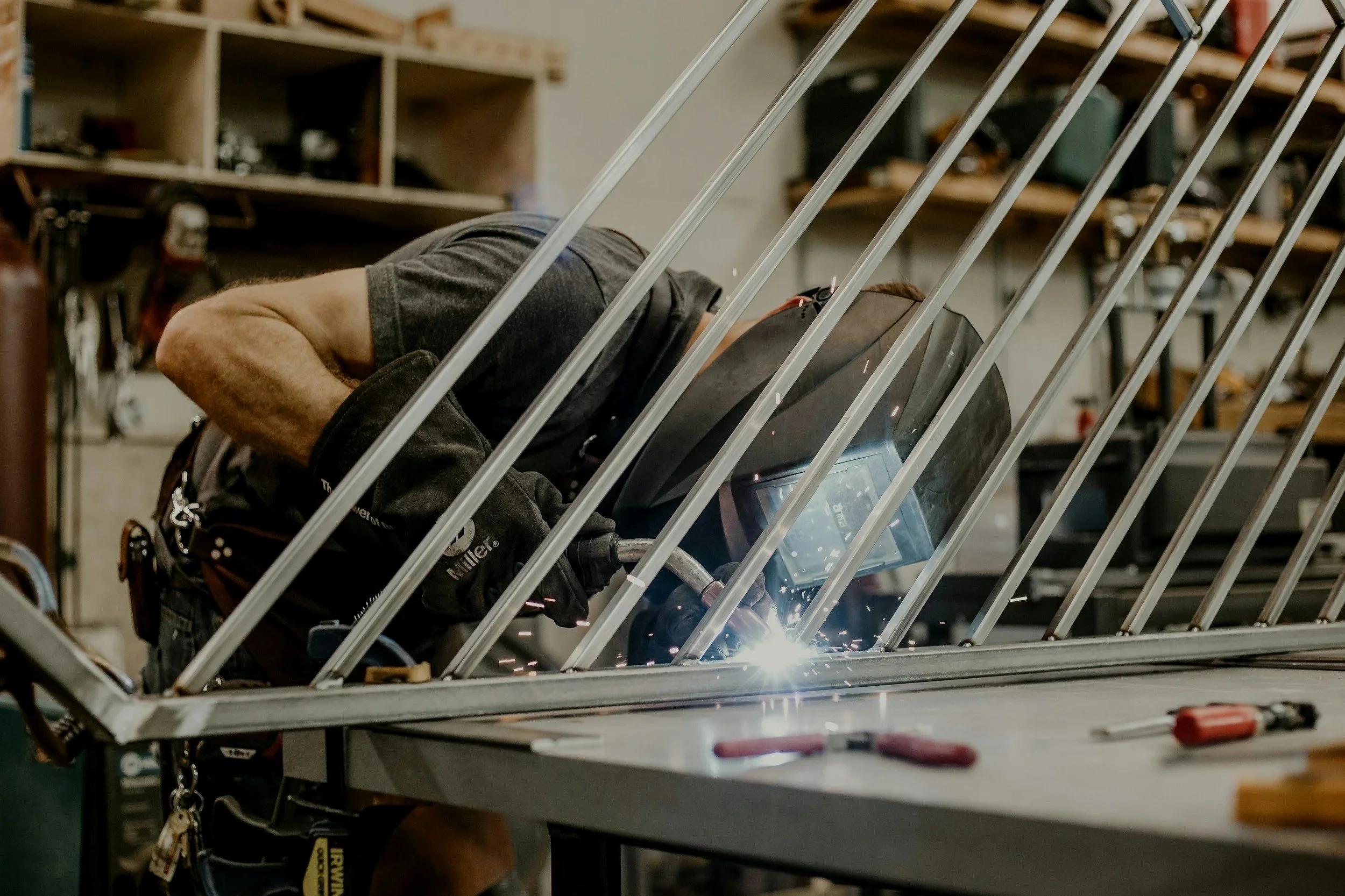 A person welding metal rods together inside a workshop. Sparks fly from the welding process, and the welder wears a protective helmet, gloves, and a dark shirt. Workbenches and tools are visible in the background. creating custom fabrication project