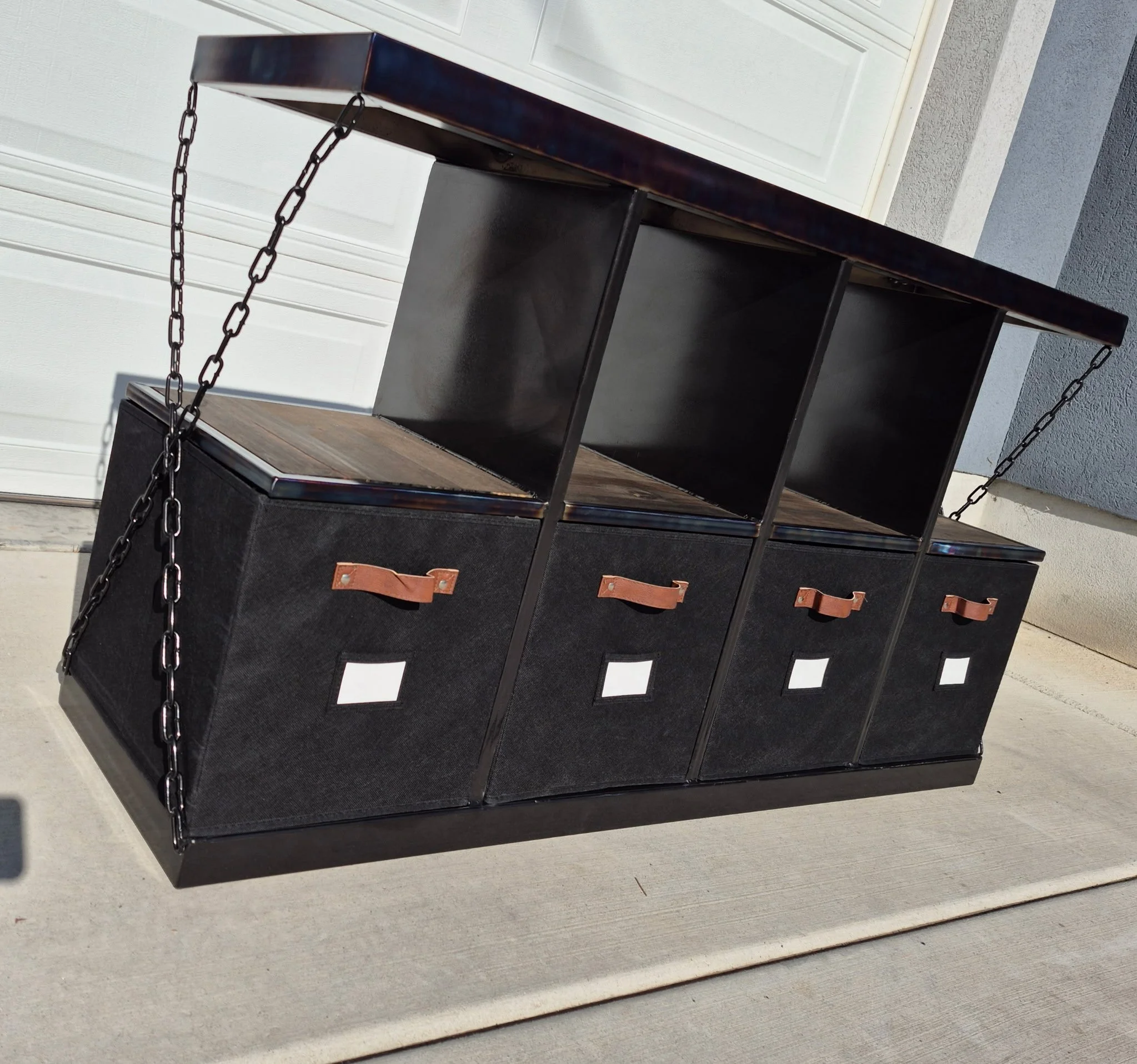 Industrial-style custom metal storage bench featuring a sleek black steel frame with a heat-treated patina finish, paired with a rich wood top and reinforced shelving. Designed with four fabric storage bins, leather pull handles, and label holders fo