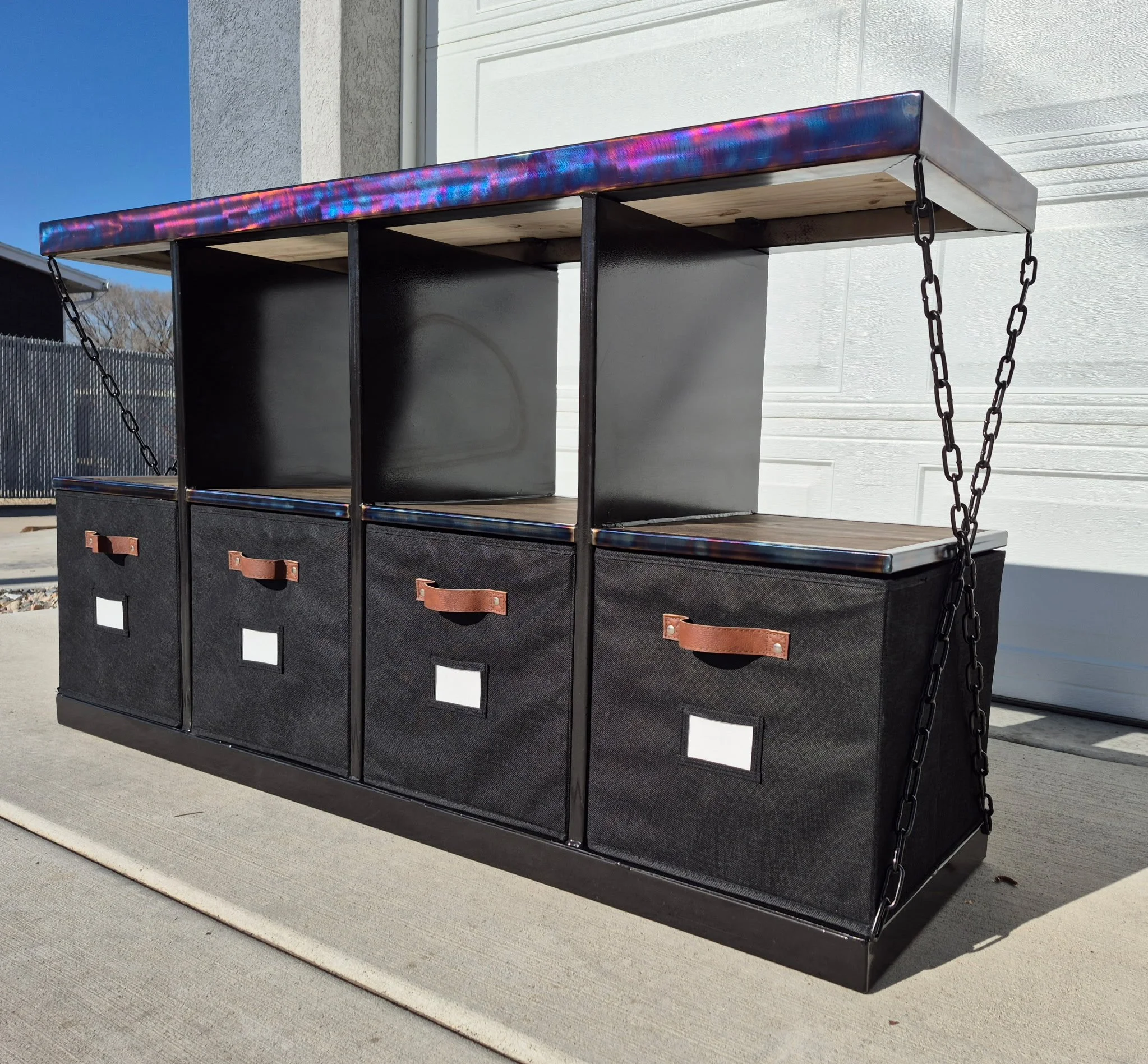 Industrial-style custom metal storage bench featuring a sleek black steel frame with a heat-treated patina finish, paired with a rich wood top and reinforced shelving. Designed with four fabric storage bins, leather pull handles, and label holders fo