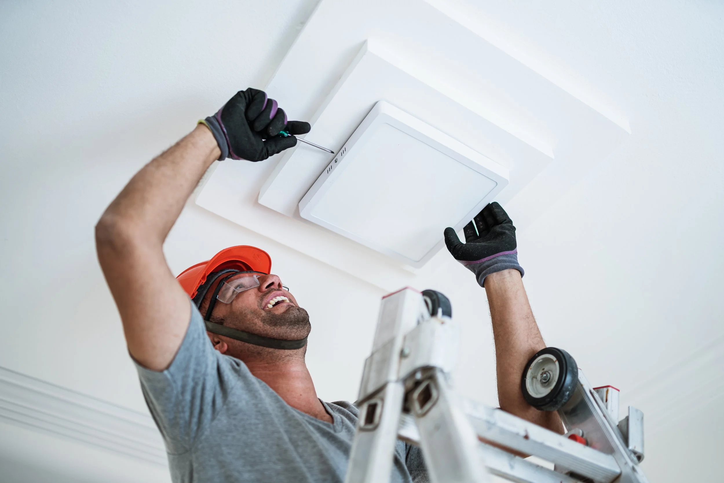 A man wearing a safety helmet, goggles, and gloves on a ladder installs a ceiling vent or light fixture in a white ceiling with a screwdriver.