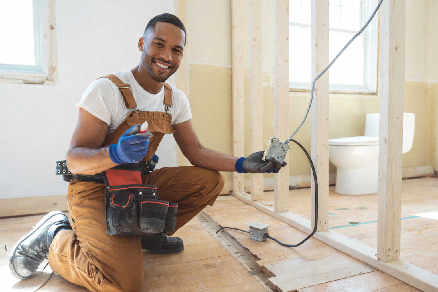 A smiling man kneeling on a wooden floor, wearing construction gloves, a tool pouch, and work overalls. He is holding electrical wiring and a switch box, working on wiring in a room with exposed wooden studs and a toilet visible in the background.