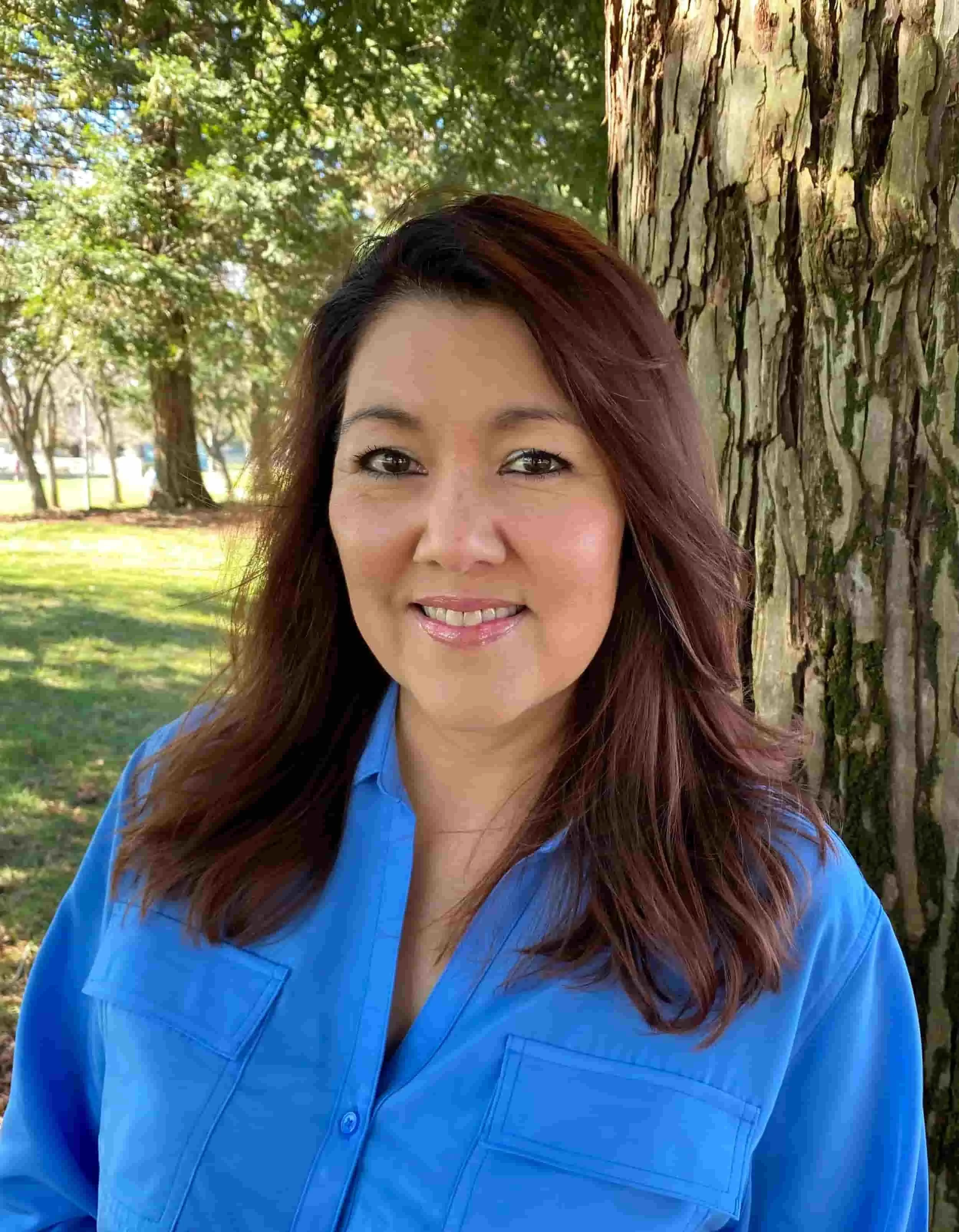 Michelle Grady stands outdoors beside a tree, smiling gently while wearing a blue blouse, with sunlight filtering through a park setting behind her.