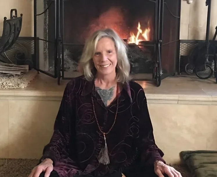 A woman with gray hair sitting in front of a lit fireplace, smiling, wearing a dark velvet top and a beaded necklace.
