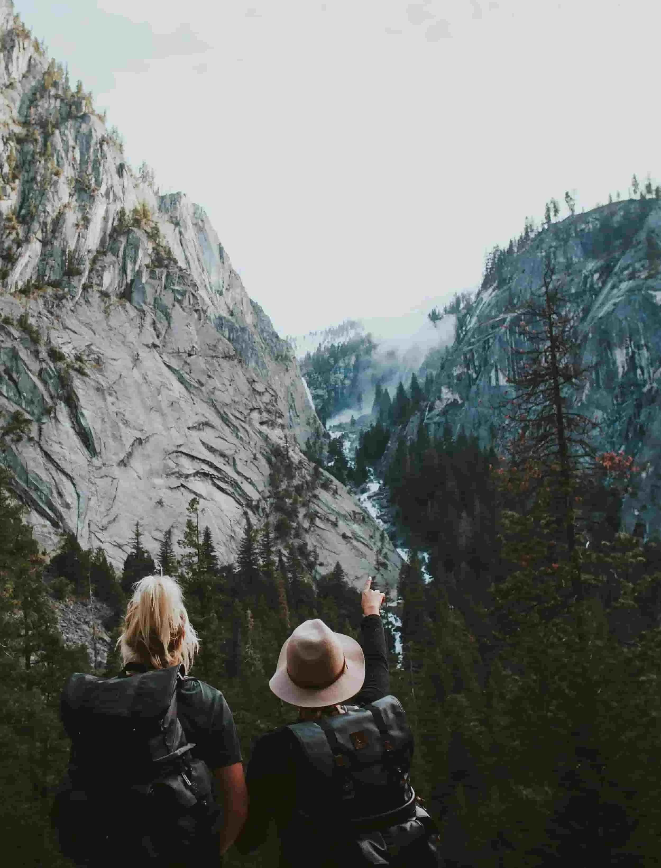 Two people with backpacks stand in forested valley looking toward steep granite cliffs and waterfall, one pointing ahead. Scene reflects shared direction curiosity and perspective often emphasized in internal family systems and ifs coaching.