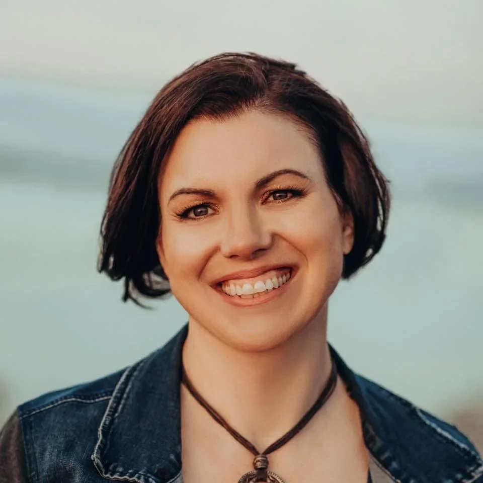 A smiling woman with short dark hair, wearing a dark denim jacket and a black necklace, outdoors with a blurred natural background.