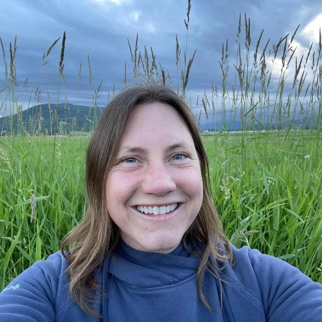 A woman smiling in a field of tall green grass with mountains and a cloudy sky in the background.