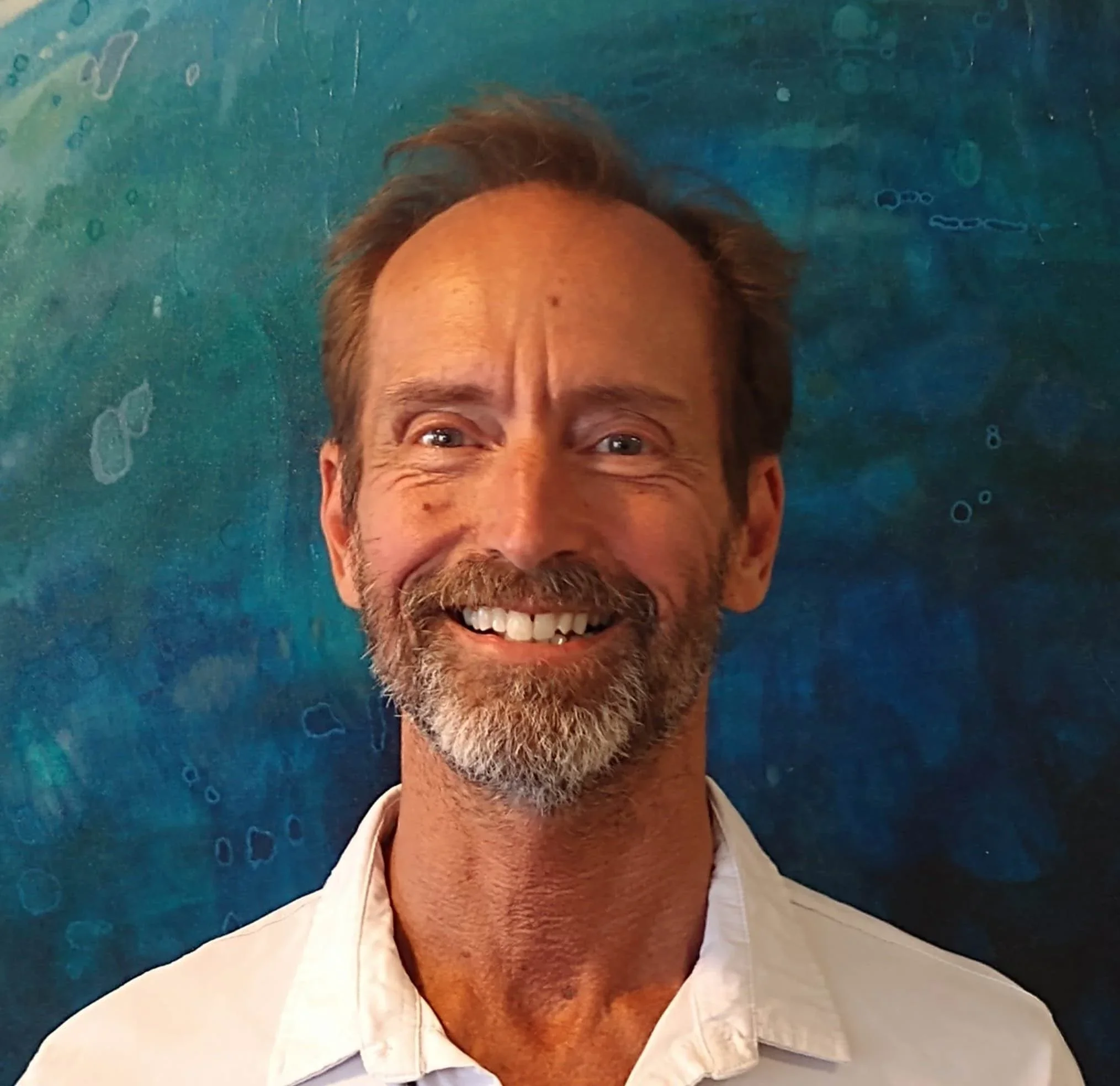 A smiling middle-aged man with a beard and mustache, wearing a white shirt, standing in front of an abstract blue and green background.