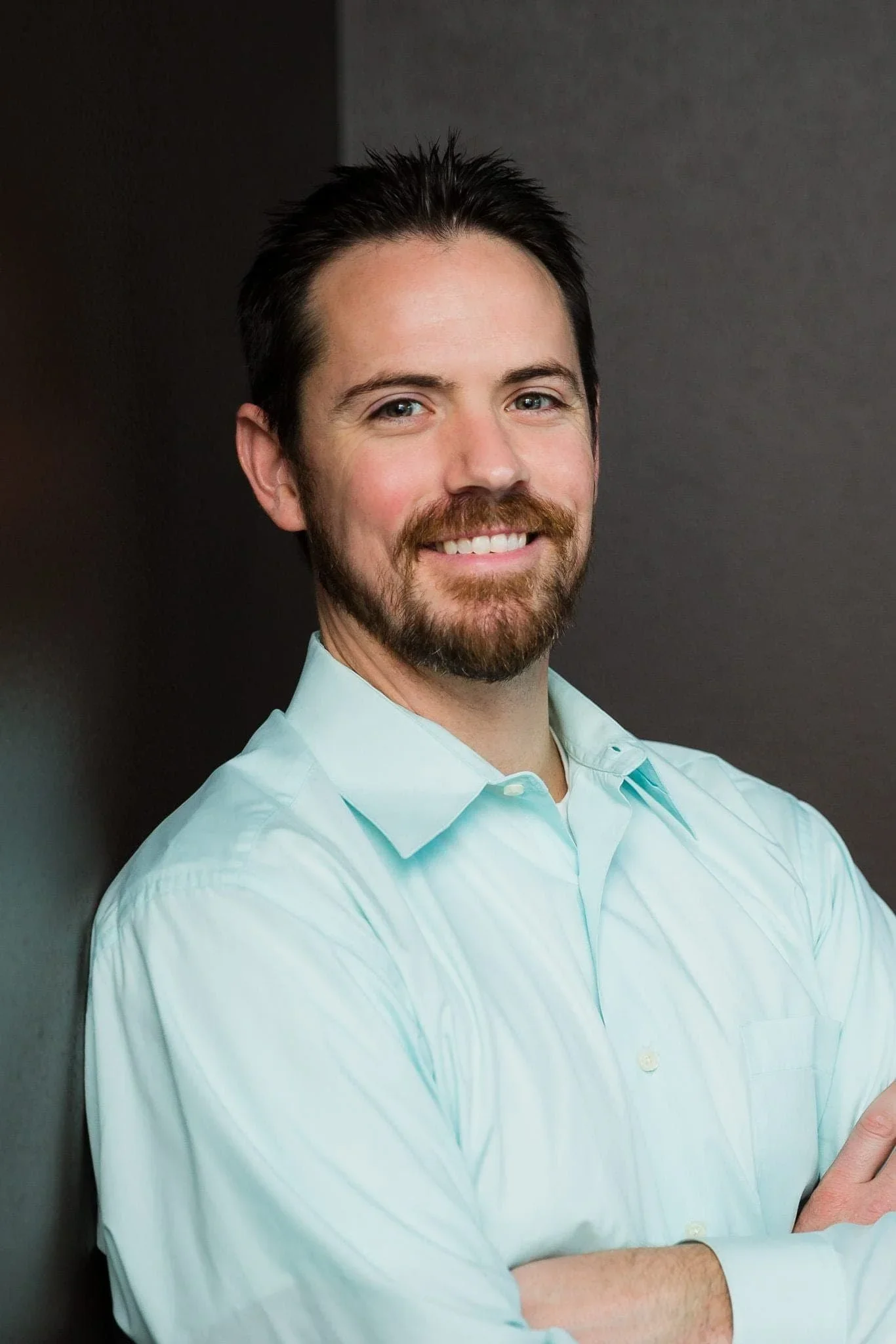 A man with dark hair, a beard, and blue eyes smiling, wearing a light blue button-up shirt, standing against a dark background.
