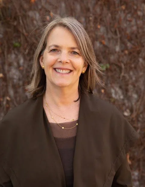 A smiling woman with shoulder-length gray hair outdoors in front of a background of brown leaves.