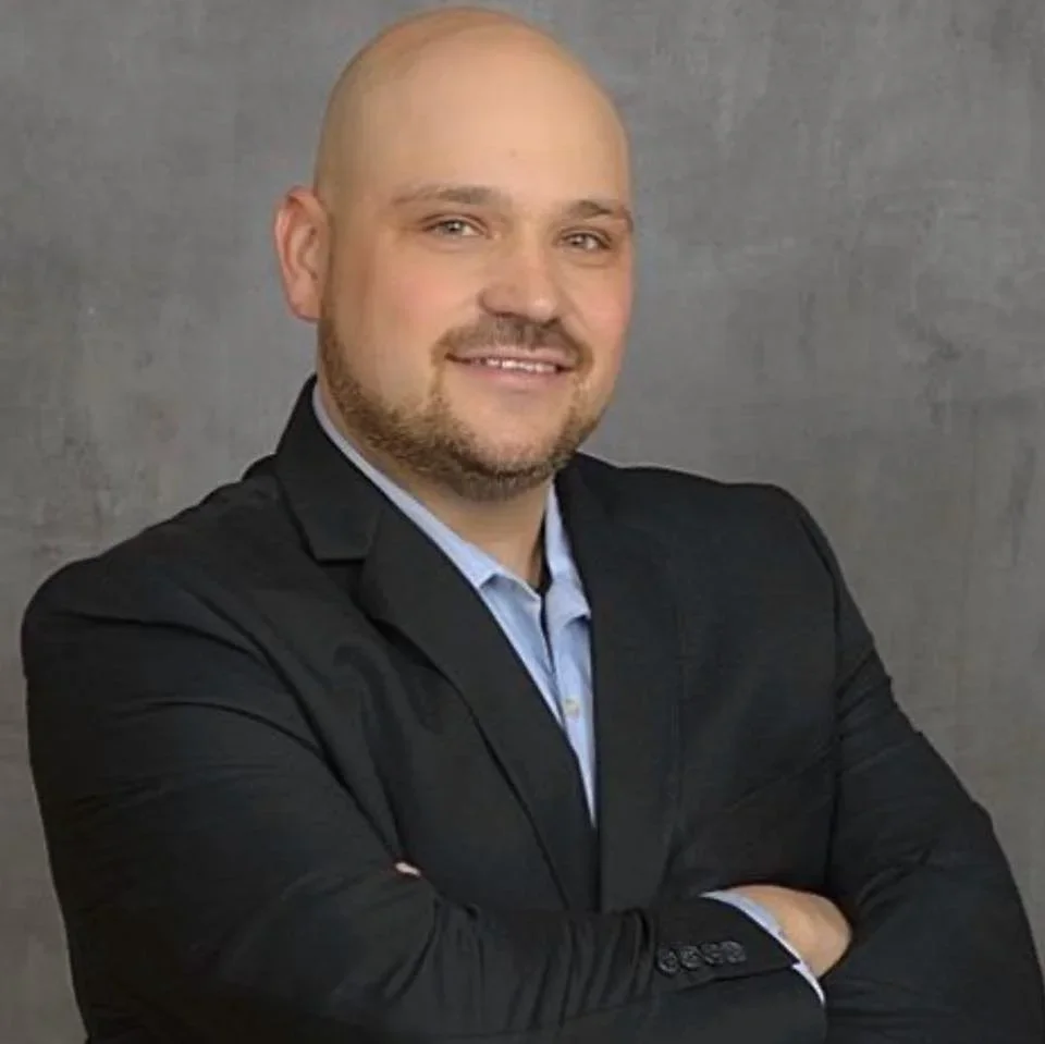 A bald man with a beard smiling, wearing a black suit jacket over a light blue dress shirt, standing with arms crossed against a grey background.