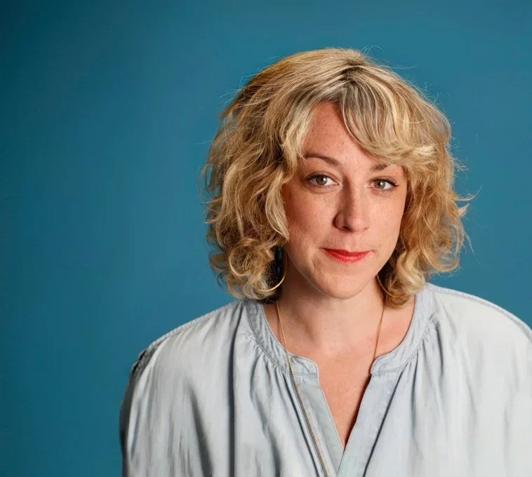 A woman with curly blonde hair wearing a light-colored blouse against a blue background.