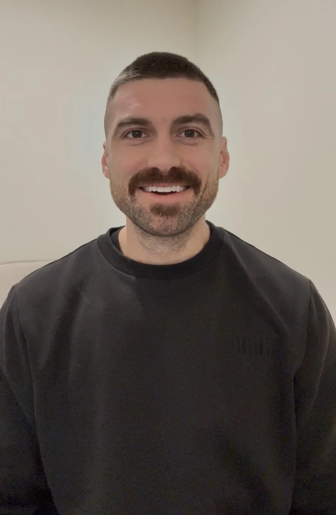 Portrait of a smiling man with a beard and short dark hair, wearing a black sweatshirt, standing against a plain white wall.