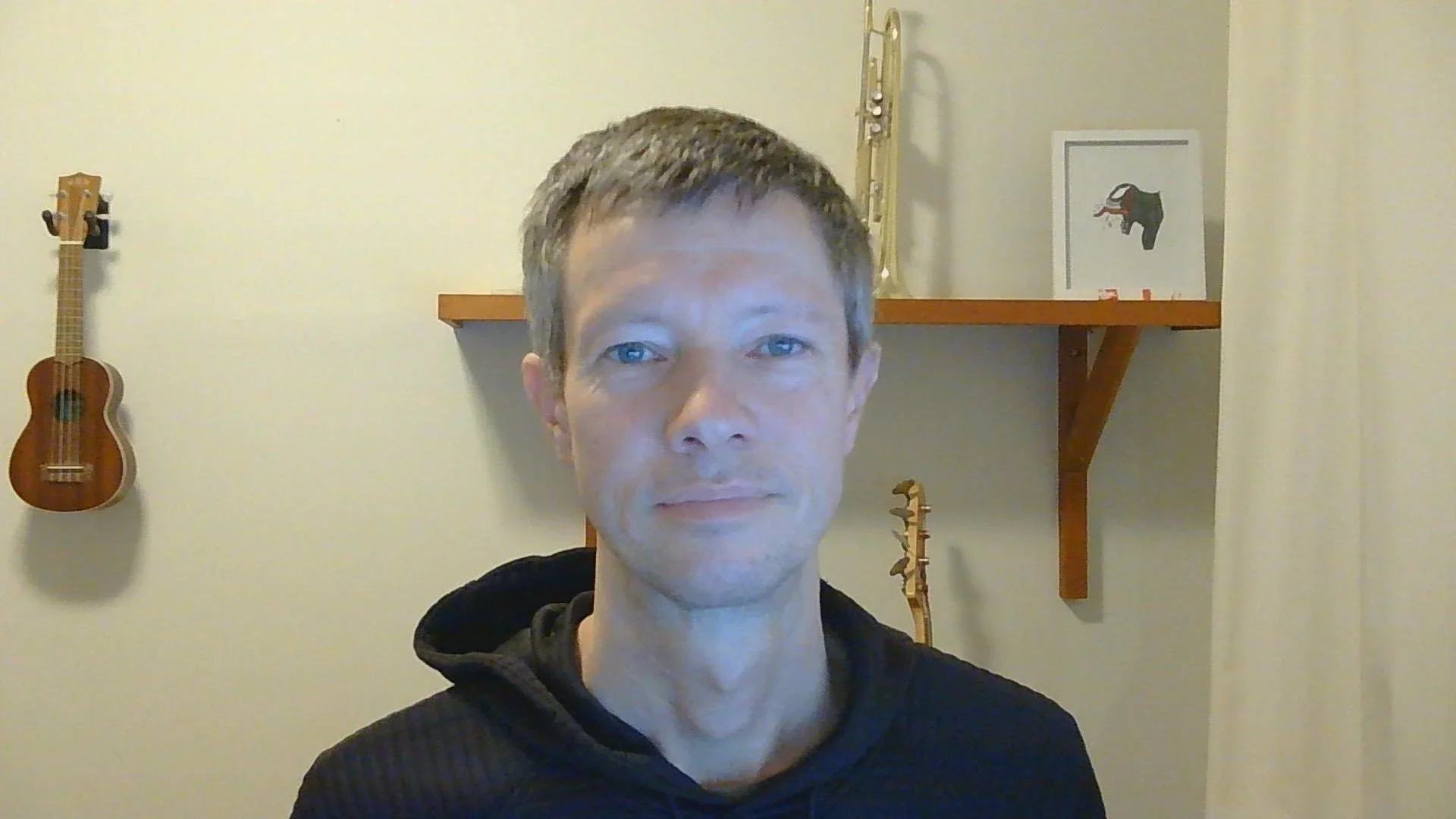 A man with light brown hair and blue eyes taking a selfie in a room with beige walls. In the background, there is a small wooden shelf with a framed art print of a black elephant with a red trunk and tusk, a small guitar model, and a tall wooden sculpture.