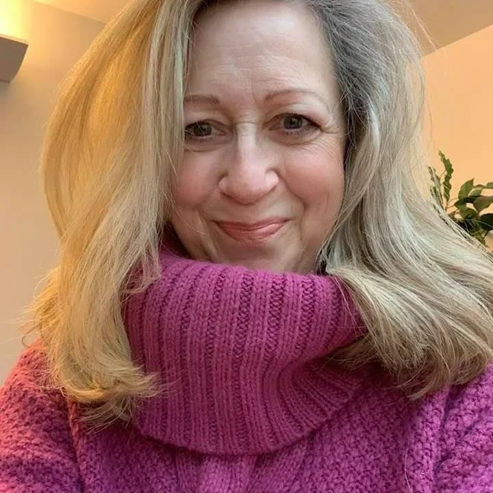A woman with shoulder-length blonde hair smiling, wearing a pink knitted sweater with a high collar.
