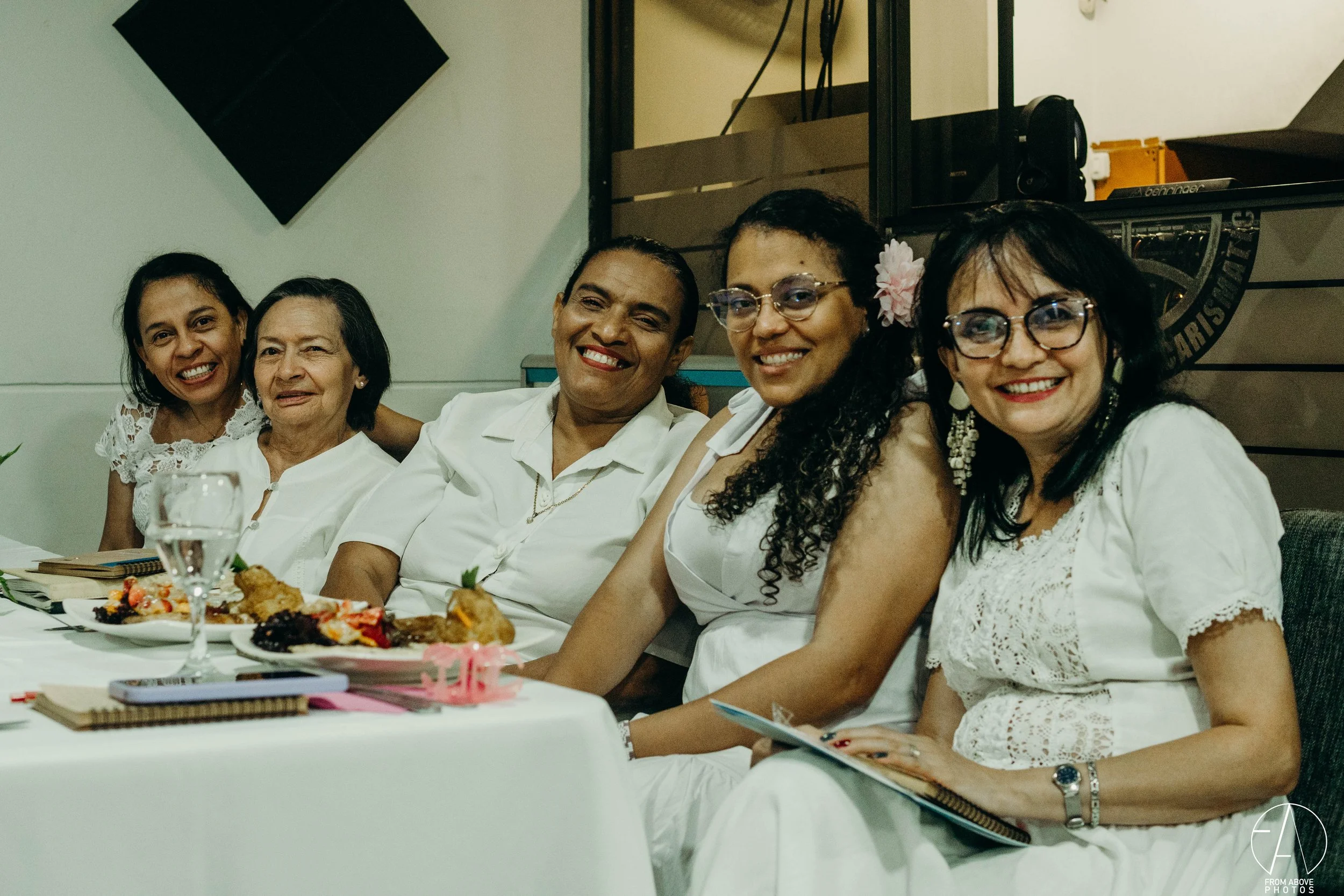 Grupo de seis mujeres sentadas en una mesa, sonriendo, vestidas con ropa blanca, en un ambiente de celebración.