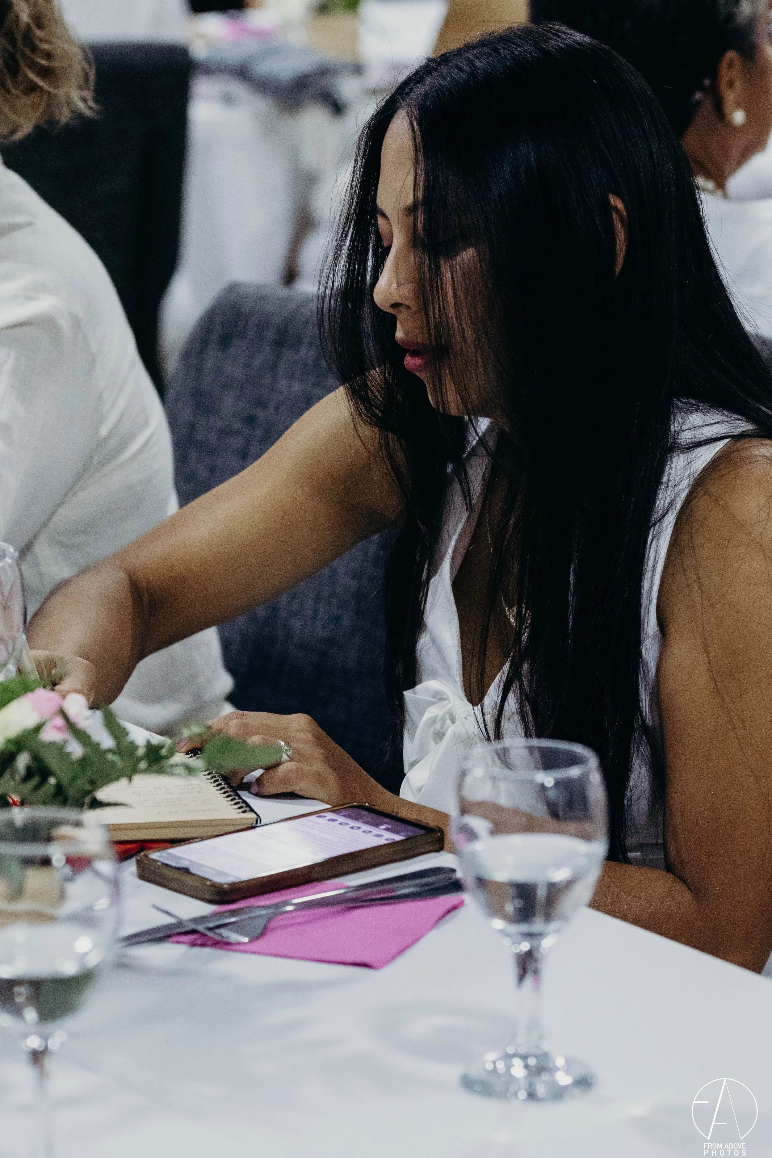 Mujer de cabello largo y oscuro en una reunión o evento, sentada en una mesa con una libreta, teléfono y vasos con agua