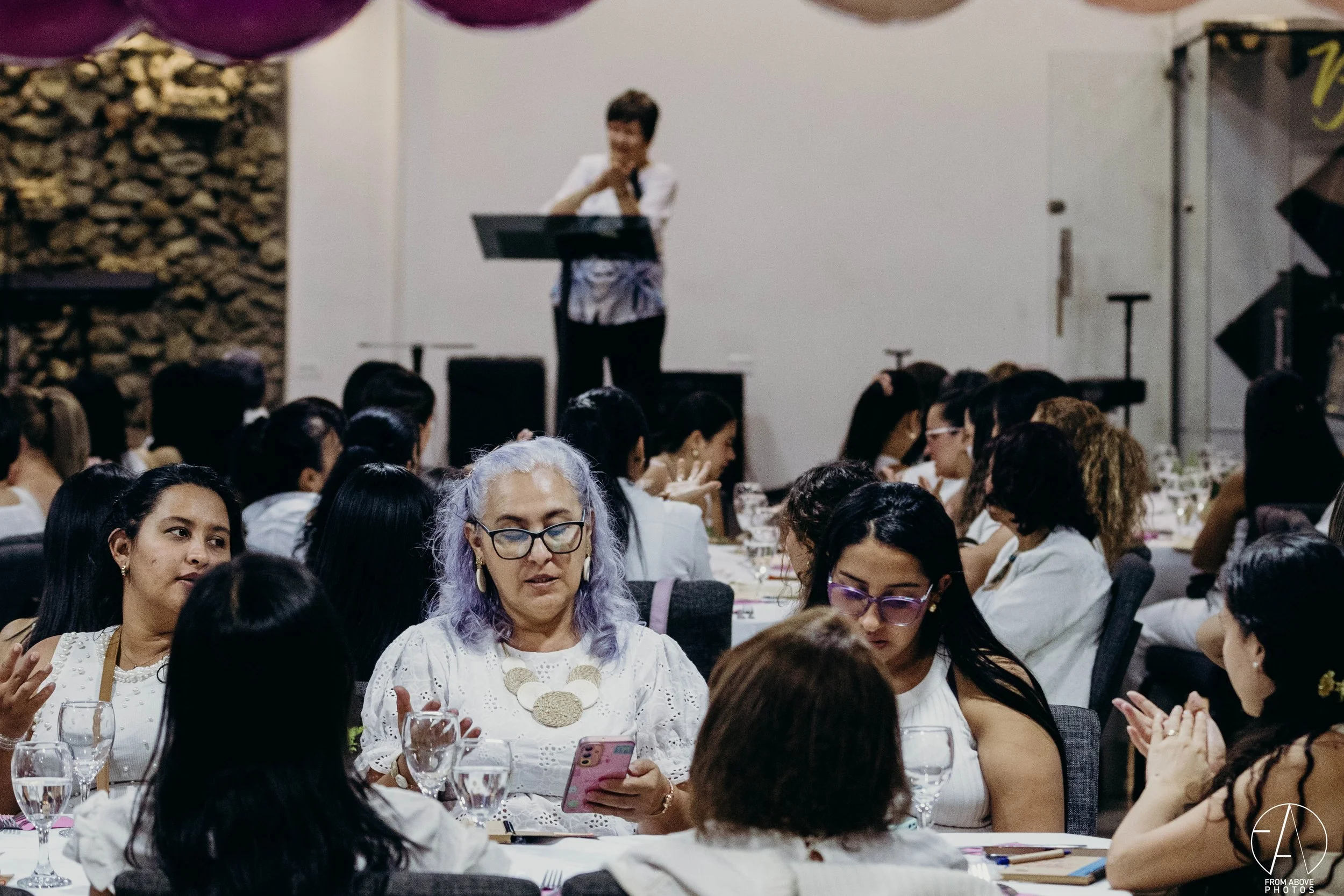 Mujeres en una reunión o evento sentadas alrededor de mesas, con una mujer en el escenario en el fondo.