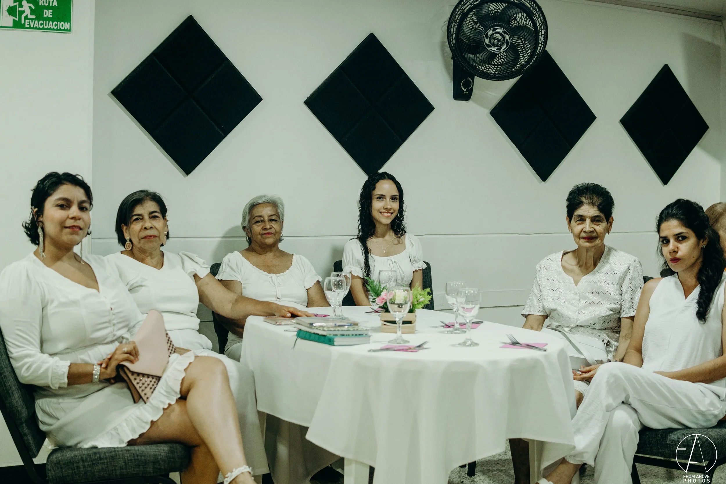 Grupo de siete mujeres sentadas alrededor de una mesa en un salón, algunas con ropa blanca, con vasos de agua y un pequeño arreglo floral en la mesa.