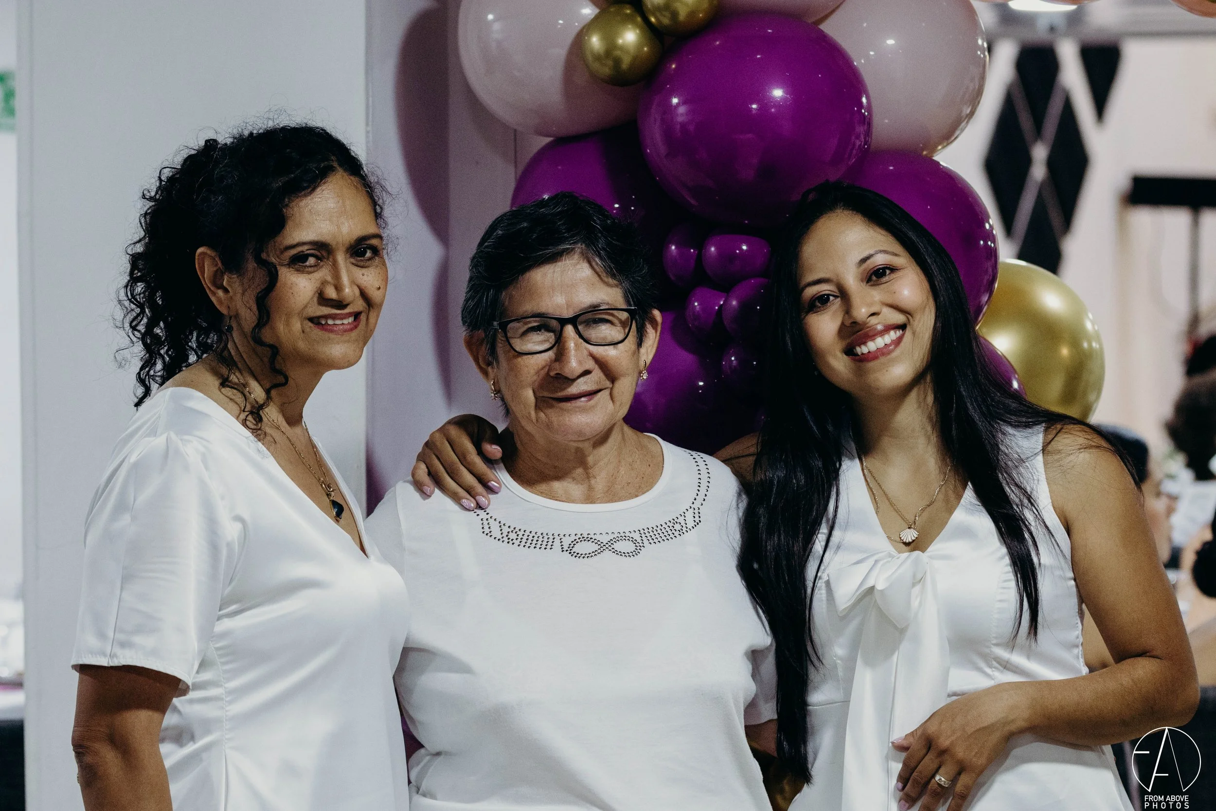 Tres mujeres posando juntas en una celebración con globos morados y dorados.