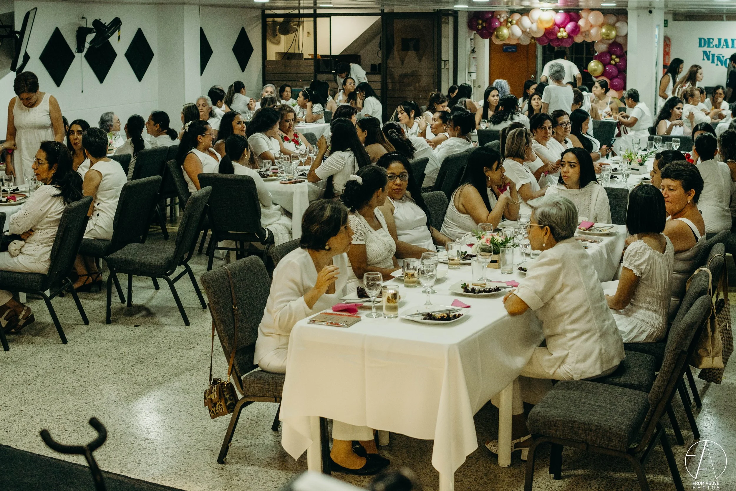 Varios grupos de mujeres sentadas en mesas decoradas con manteles blancos, en un evento social. Algunas están conversando y otras están comiendo, en un ambiente festivo con globos en el fondo.