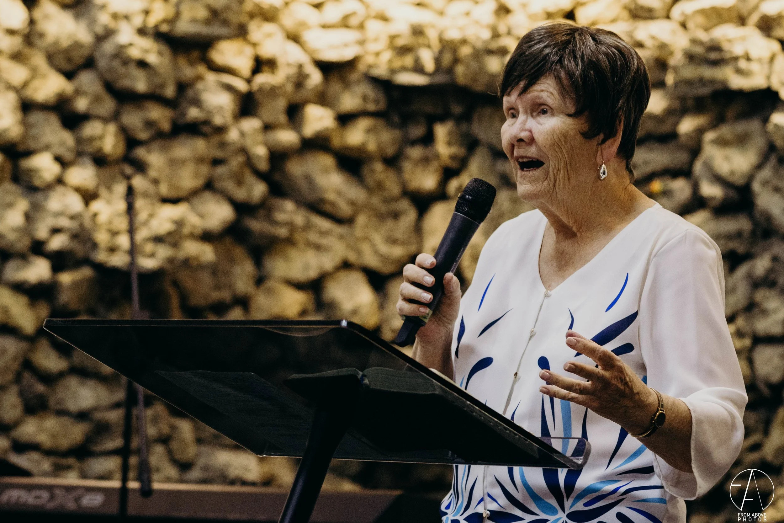 Una mujer mayor habla con un micrófono frente a una pared de piedra, en lo que parece ser una presentación o discurso.