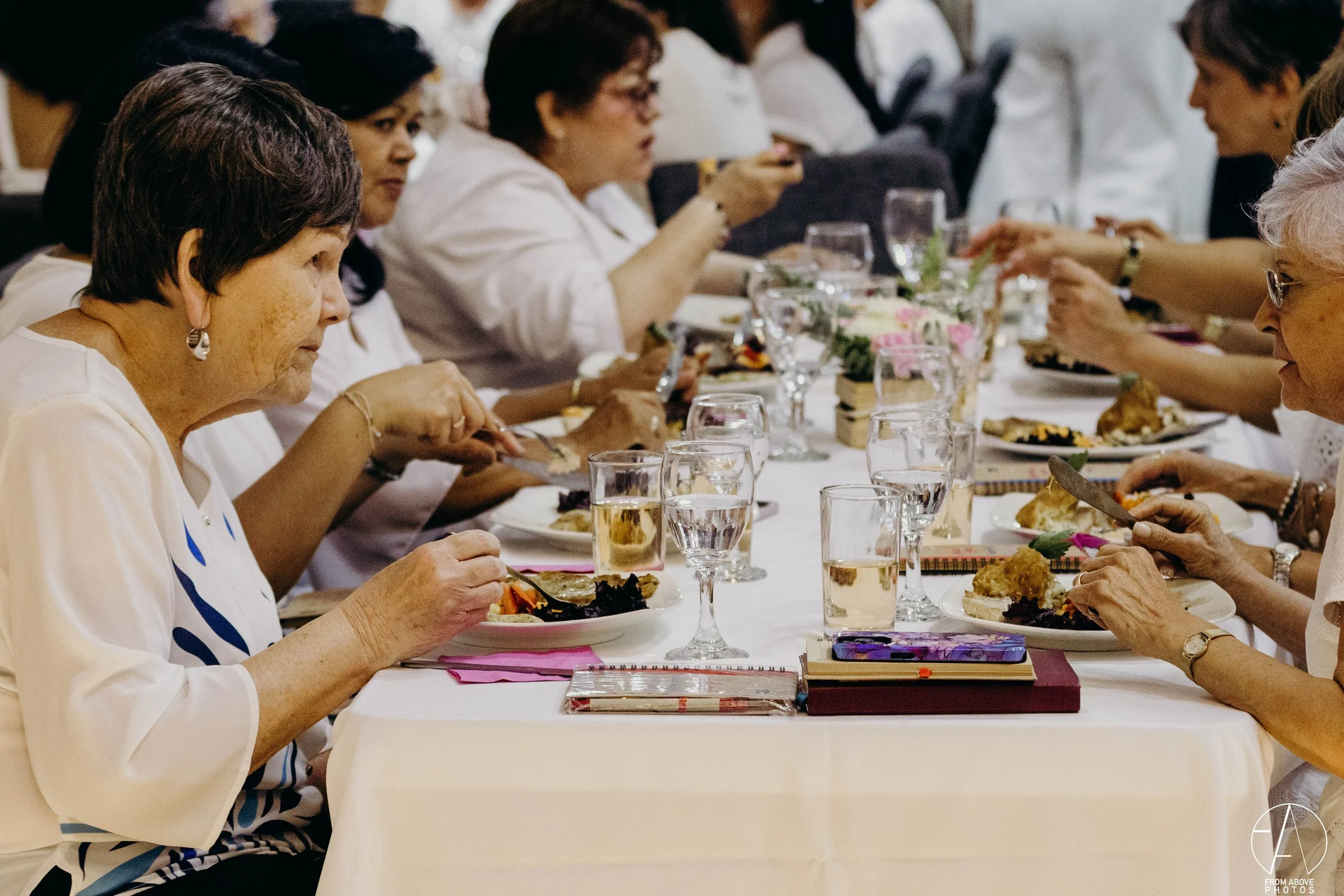 Grupo de mujeres mayores sentadas en una mesa larga, comiendo y bebiendo en un evento social, con platos de comida y vasos de agua sobre la mesa